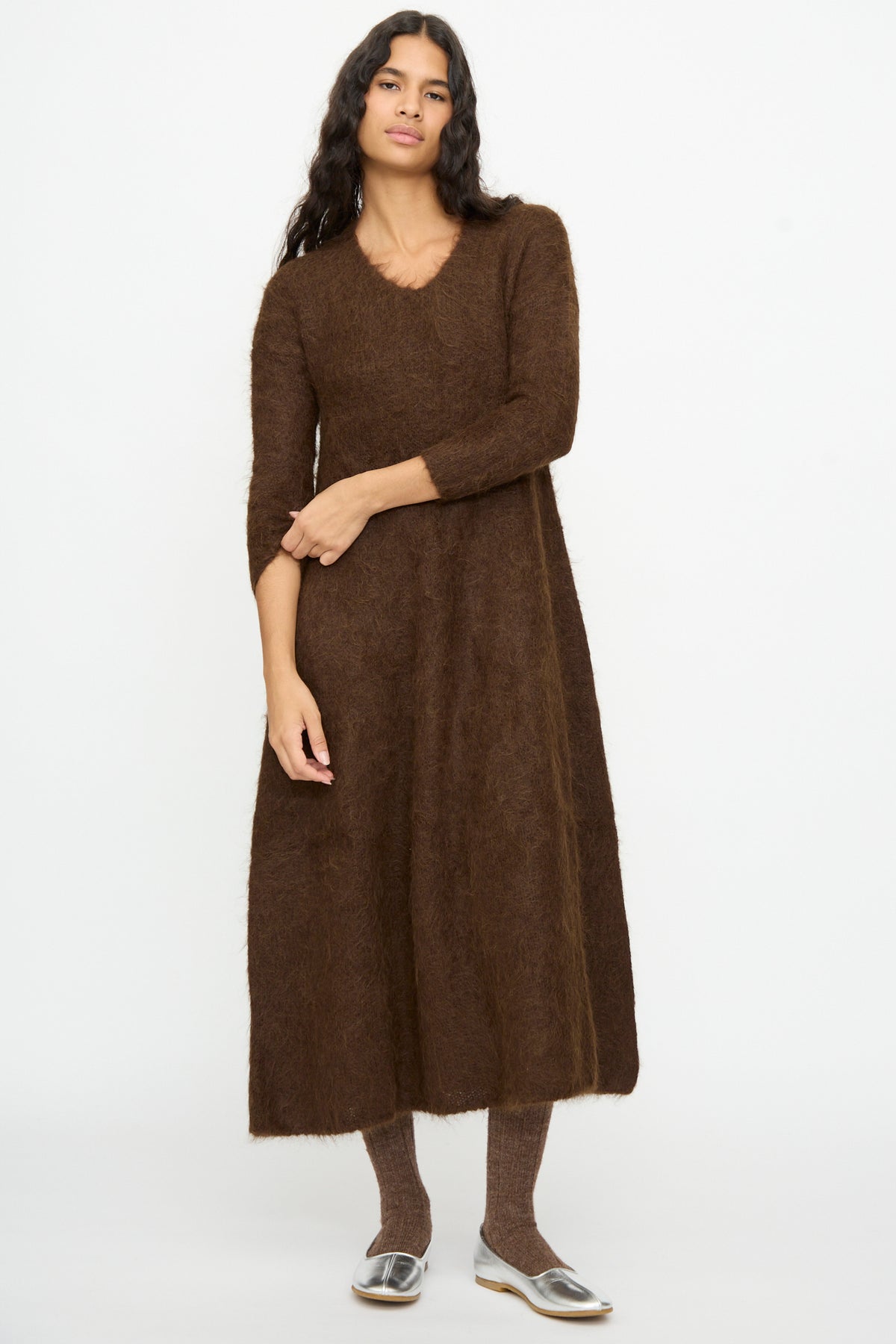 A woman wears a brown Cosmic Wonder Alpaca Knitted Evening Dress, a V-neck midi-length knit piece with long sleeves, styled with brown socks and silver flats.
