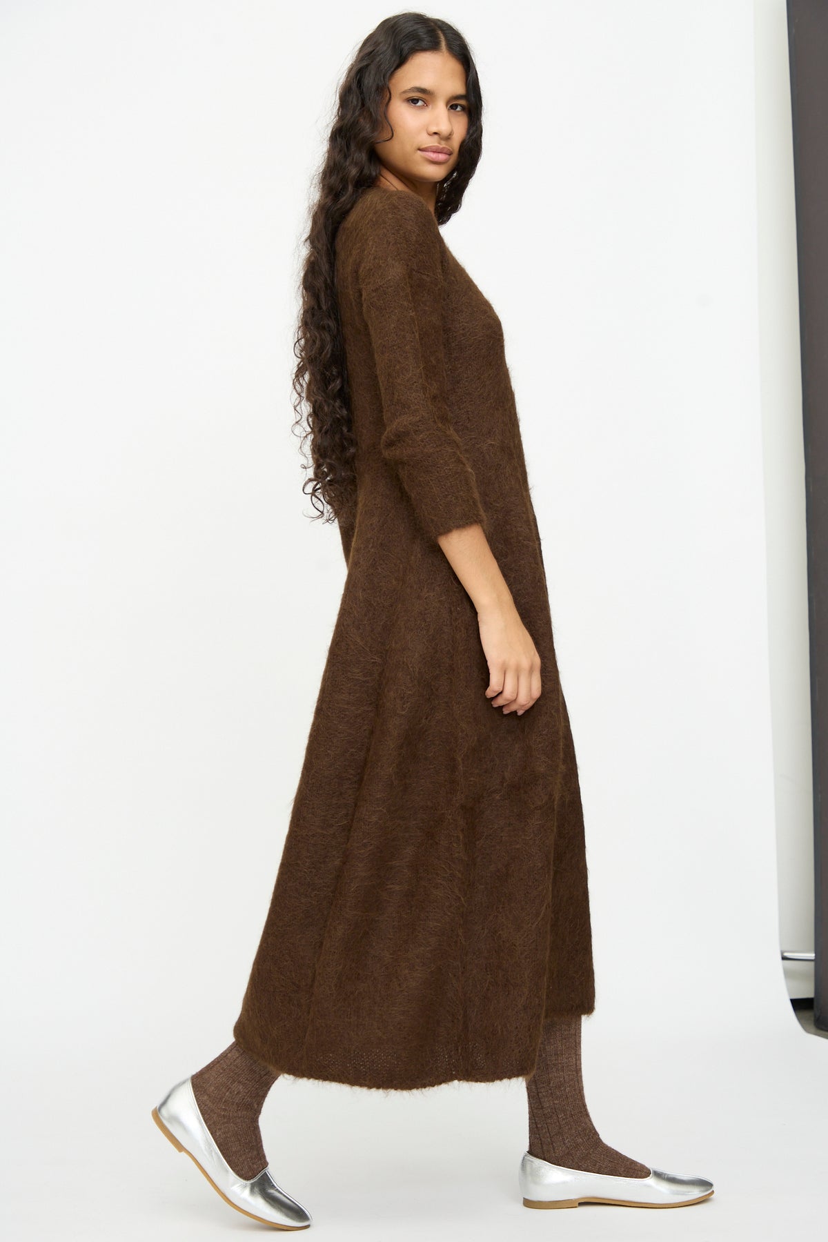 Model in a brown, full-length Cosmic Wonder Alpaca Knitted Evening Dress with long sleeves, paired with brown knit socks and silver flat shoes.