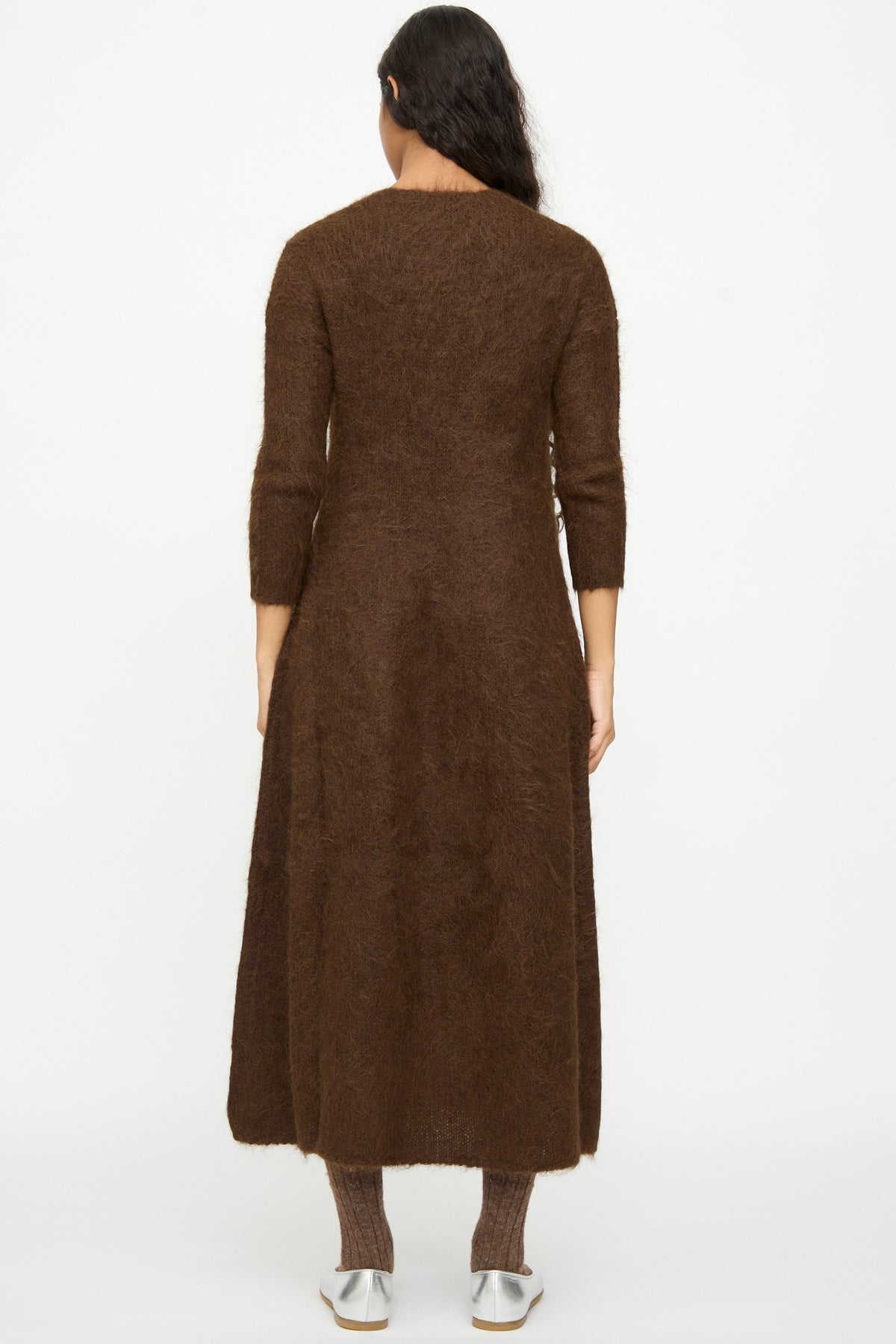 Back view of a person wearing a brown, long-sleeved, ankle-length Cosmic Wonder Alpaca Knitted Evening Dress with matching socks and silver flat shoes.