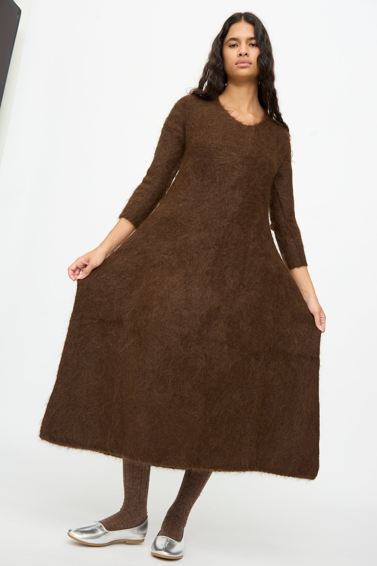 A person wearing a brown, long-sleeved Cosmic Wonder Alpaca Knitted Evening Dress with a flared skirt, brown tights, and silver flats.