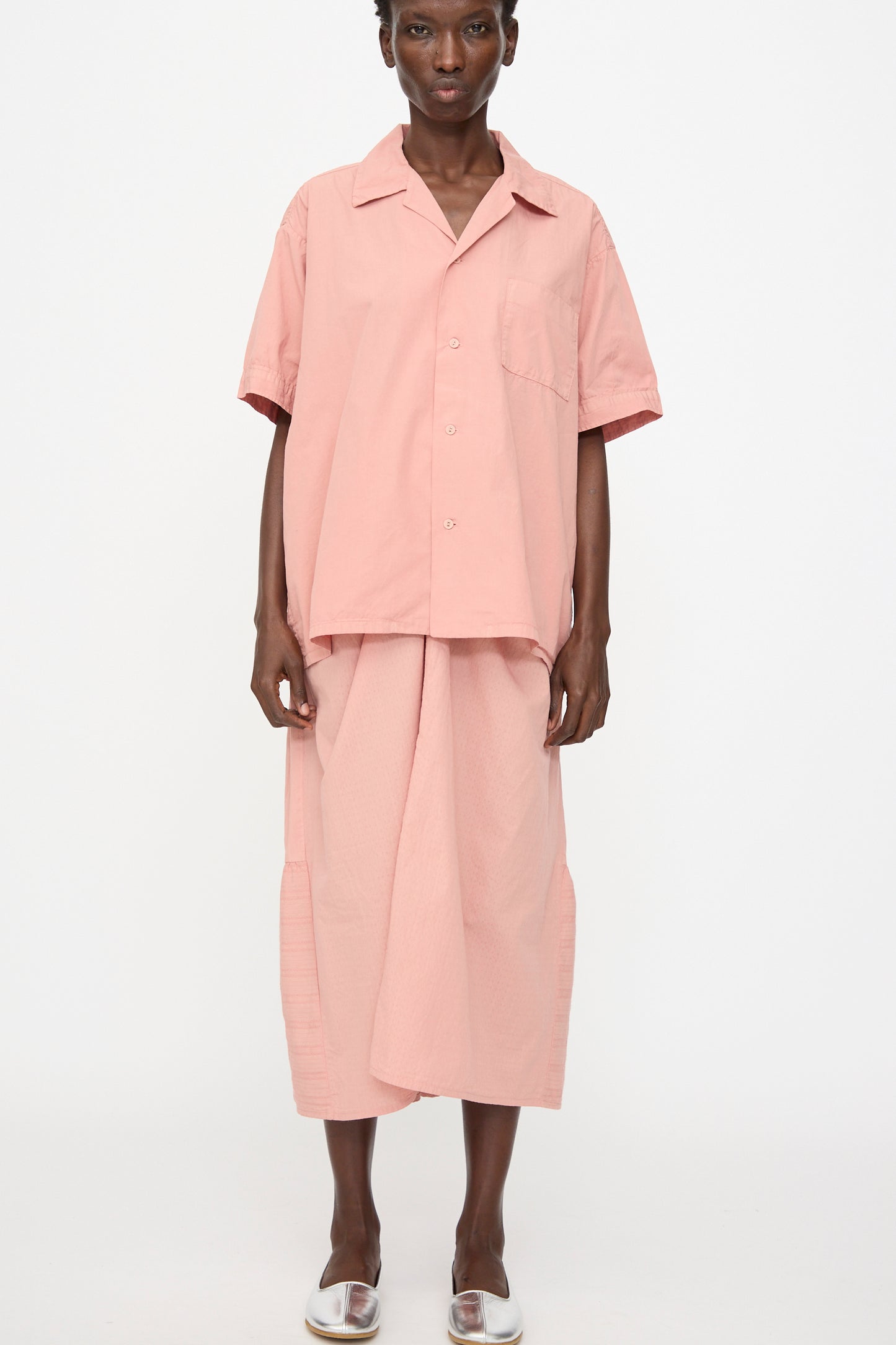 A person stands against a white background wearing the Cosmic Wonder Cotton Broadcloth Open Collar Shirt in Rose Crystal, paired with a matching midi skirt and clear open-toe shoes.