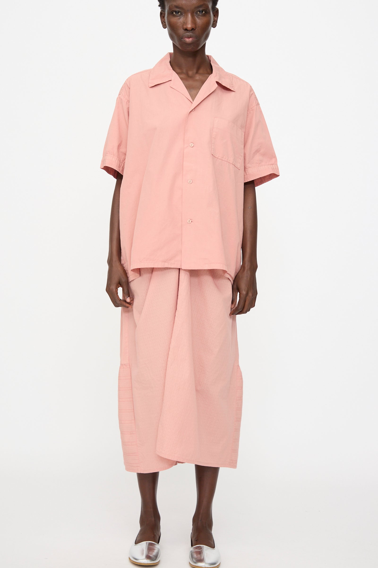 A person stands against a white background wearing the Cosmic Wonder Cotton Broadcloth Open Collar Shirt in Rose Crystal, paired with a matching midi skirt and clear open-toe shoes.