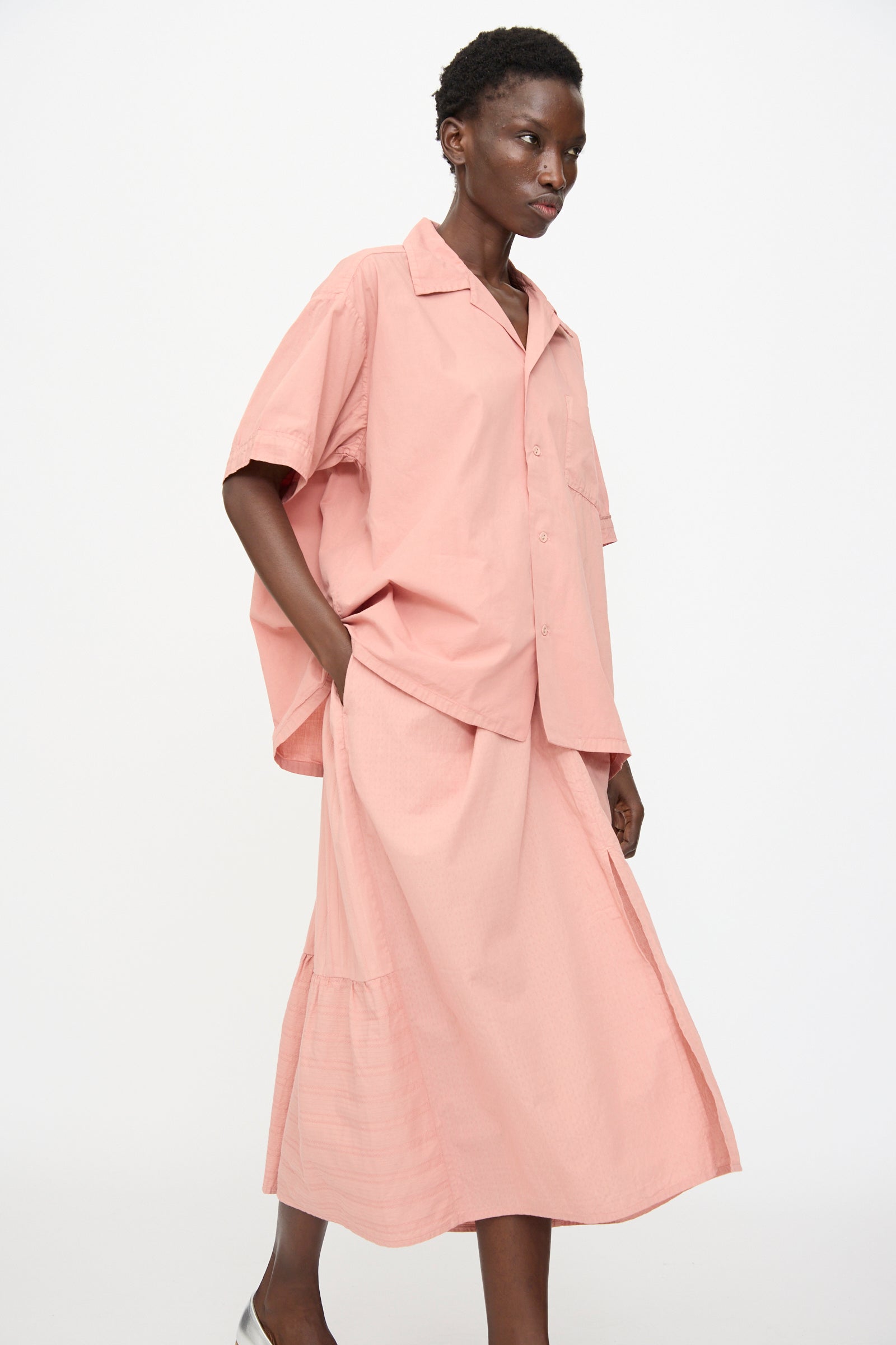 A person stands against a plain background wearing the Cosmic Wonder Cotton Broadcloth Open Collar Shirt in Rose Crystal, styled with a matching midi skirt and silver shoes.