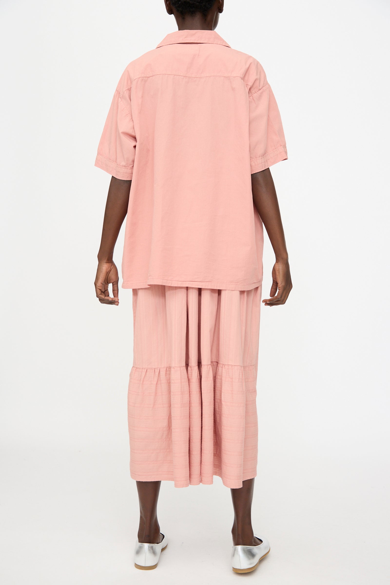 A person stands facing away against a white background, wearing Cosmic Wonder's Cotton Broadcloth Open Collar Shirt in Rose Crystal and a matching pink skirt with white slip-on shoes.