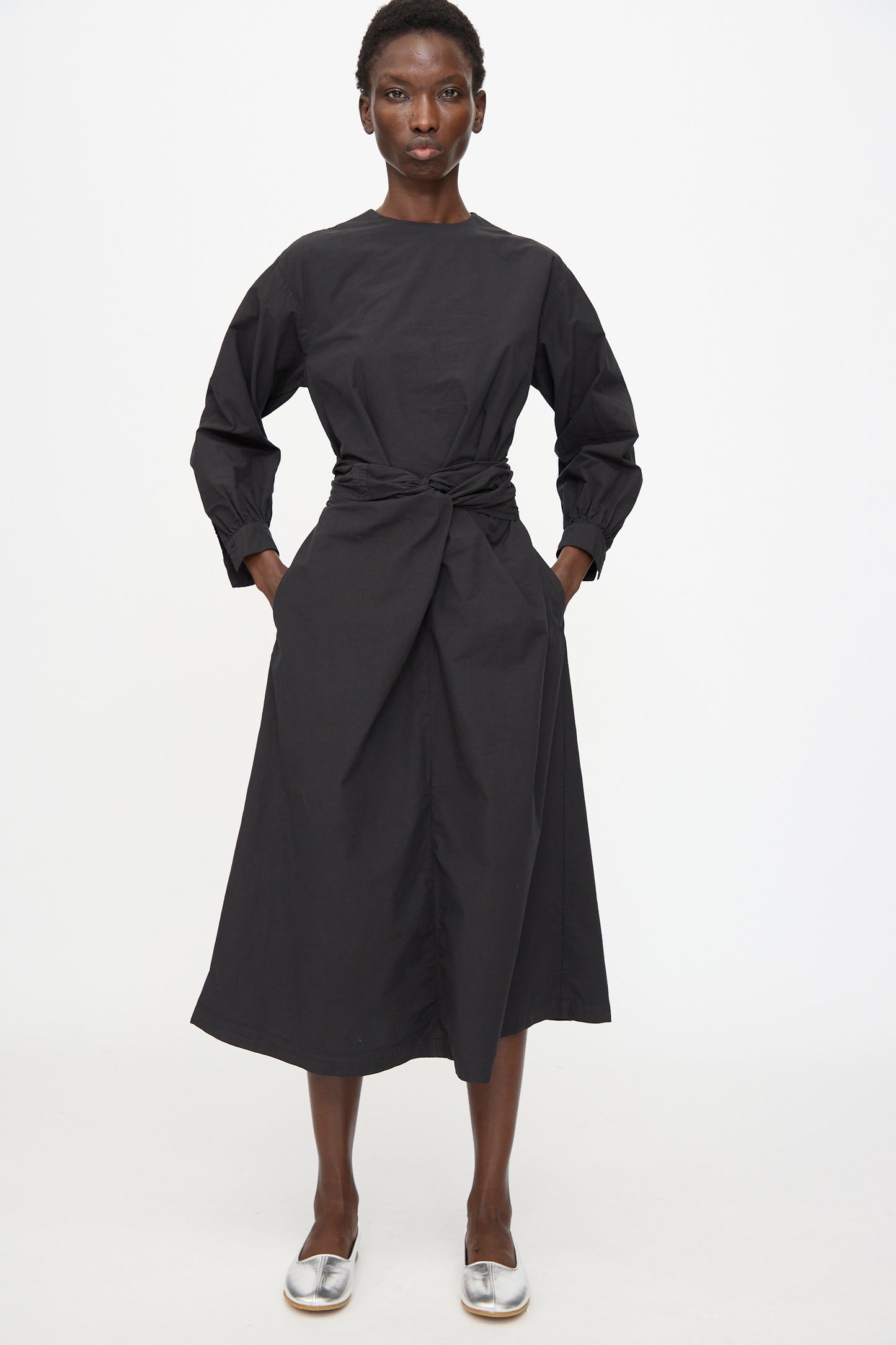 A person wears the Cosmic Wonder Cotton Broadcloth Wrapped Dress in Black, featuring long sleeves and a waist tie, paired with silver flat shoes, and stands against a plain white background.