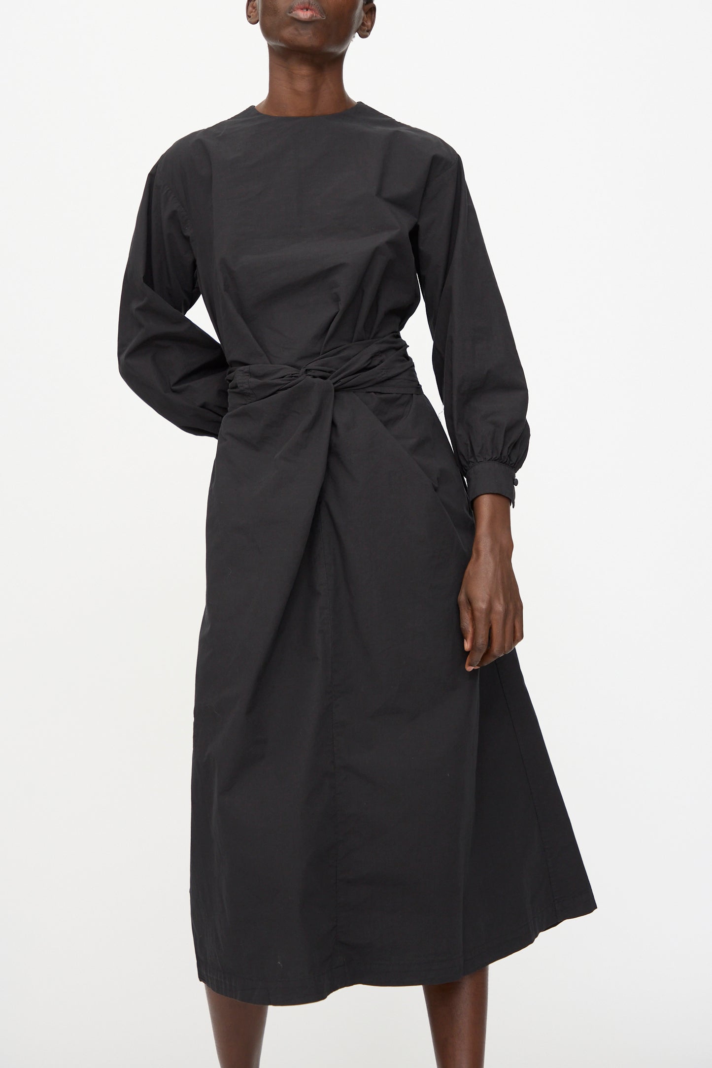 A person wearing the Cosmic Wonder Cotton Broadcloth Wrapped Dress in Black—an organic cotton, long-sleeve, ankle-length dress with a gathered waist—stands against a plain white background.