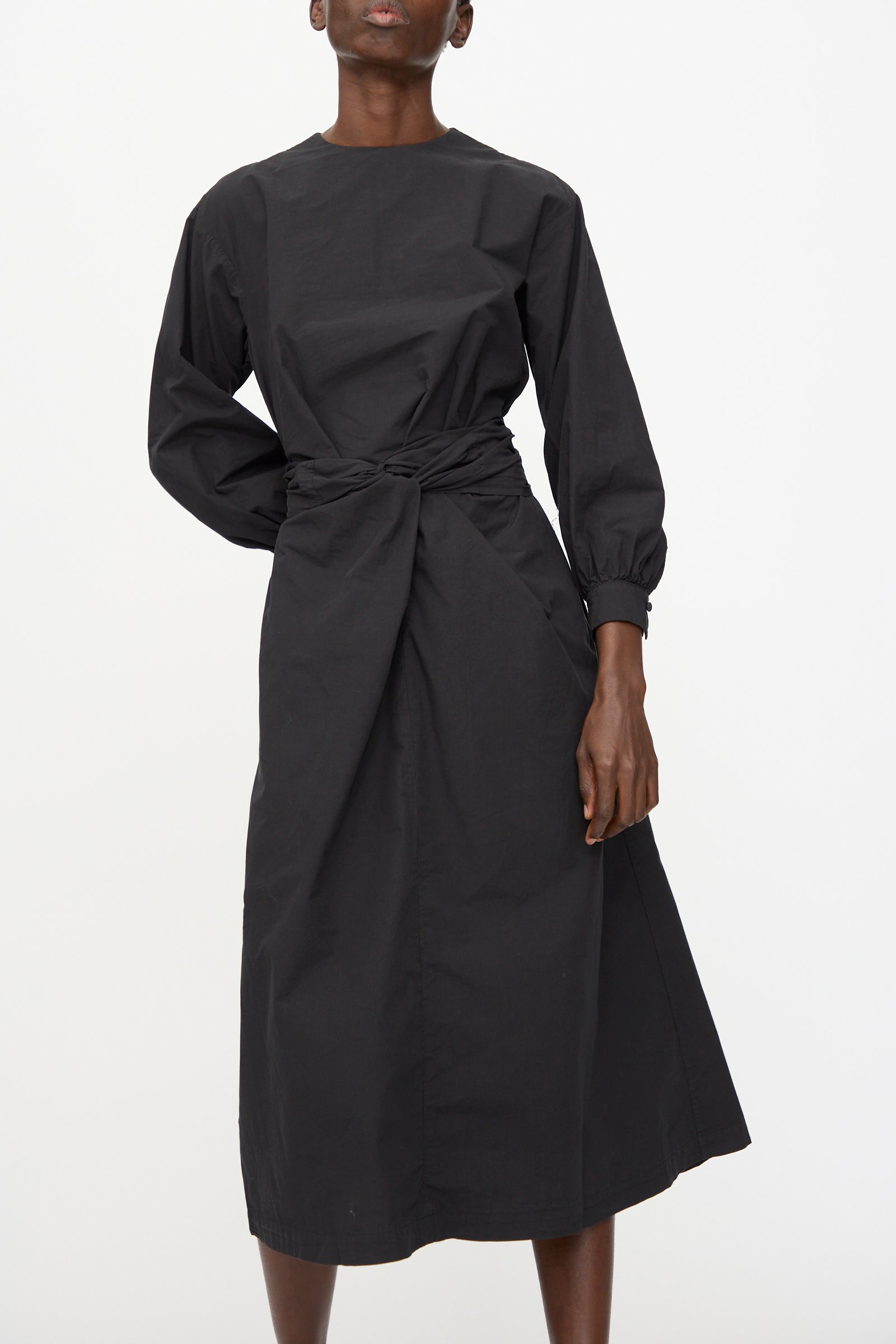 A person wearing the Cosmic Wonder Cotton Broadcloth Wrapped Dress in Black—an organic cotton, long-sleeve, ankle-length dress with a gathered waist—stands against a plain white background.