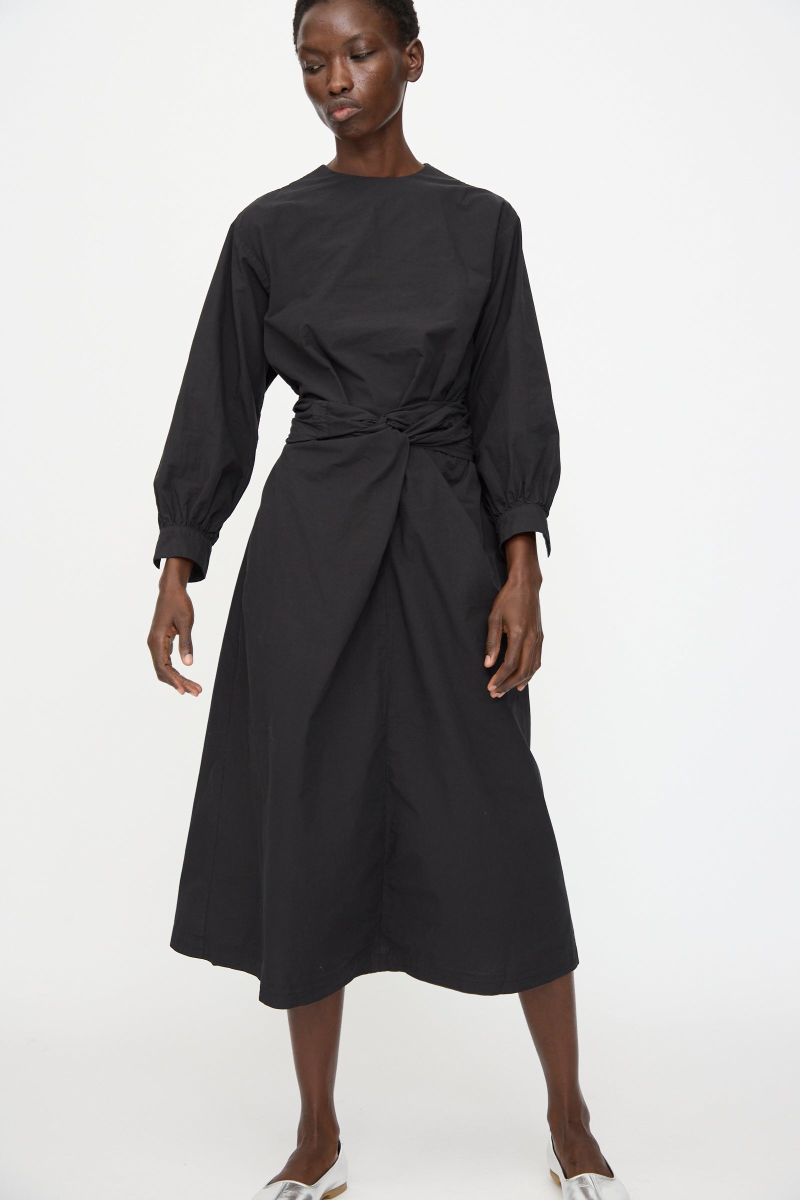 A person wears the Cosmic Wonder Cotton Broadcloth Wrapped Dress in Black, featuring long sleeves and a front knot, made from organic cotton, against a plain white background.