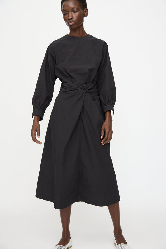 A person wears the Cosmic Wonder Cotton Broadcloth Wrapped Dress in Black, featuring long sleeves and a front knot, made from organic cotton, against a plain white background.