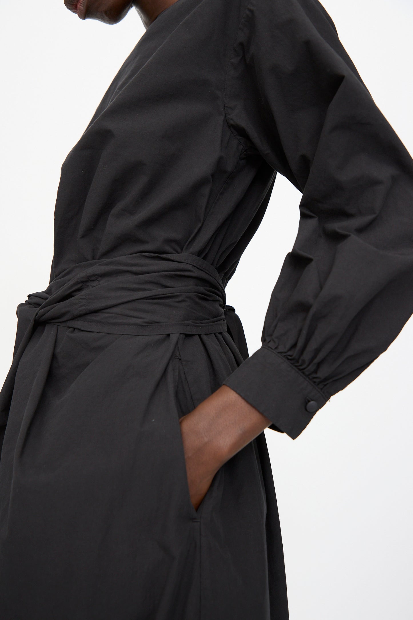 A person wears the Cosmic Wonder Cotton Broadcloth Wrapped Dress in Black, featuring long sleeves, a tied waist, and crafted from soft organic cotton, shown from shoulders to mid-thigh with one hand in the pocket.