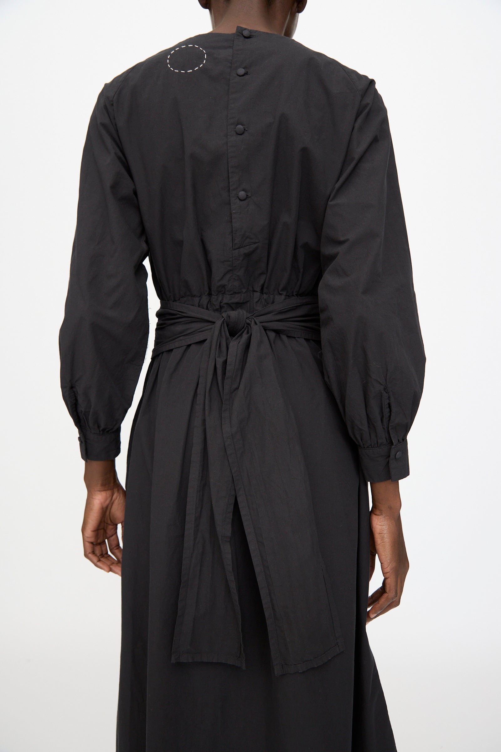 A person is seen from the back wearing the Cosmic Wonder Cotton Broadcloth Wrapped Dress in black, featuring long sleeves, a tied waist belt, and buttons along the upper back.