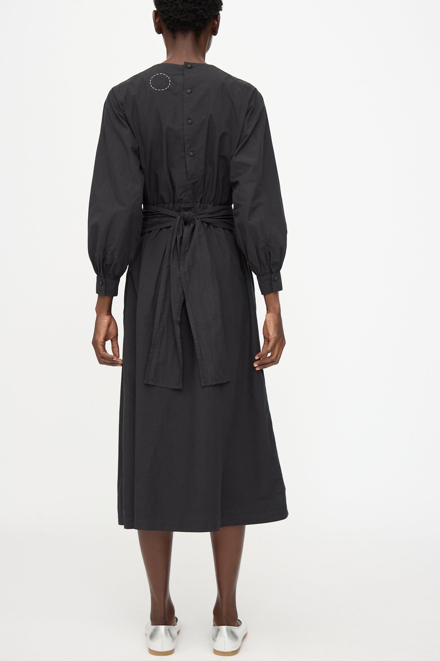 A model wears the Cosmic Wonder Cotton Broadcloth Wrapped Dress in Black, featuring long sleeves, a button-down back, and tied waist, standing against a plain white background.