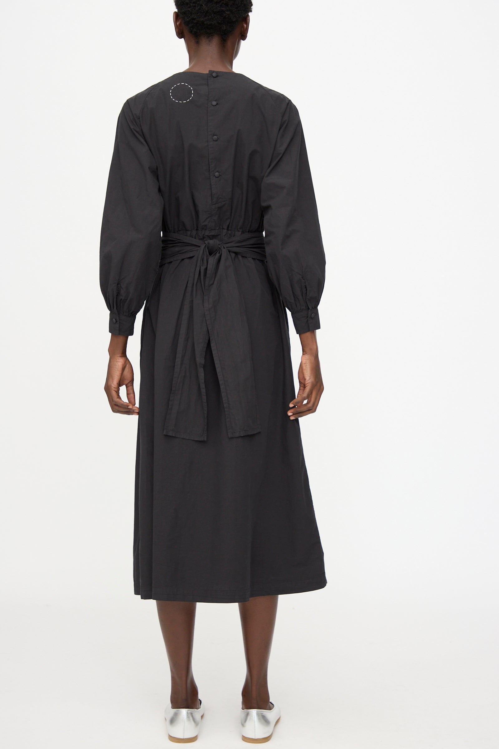 A model wears the Cosmic Wonder Cotton Broadcloth Wrapped Dress in Black, featuring long sleeves, a button-down back, and tied waist, standing against a plain white background.