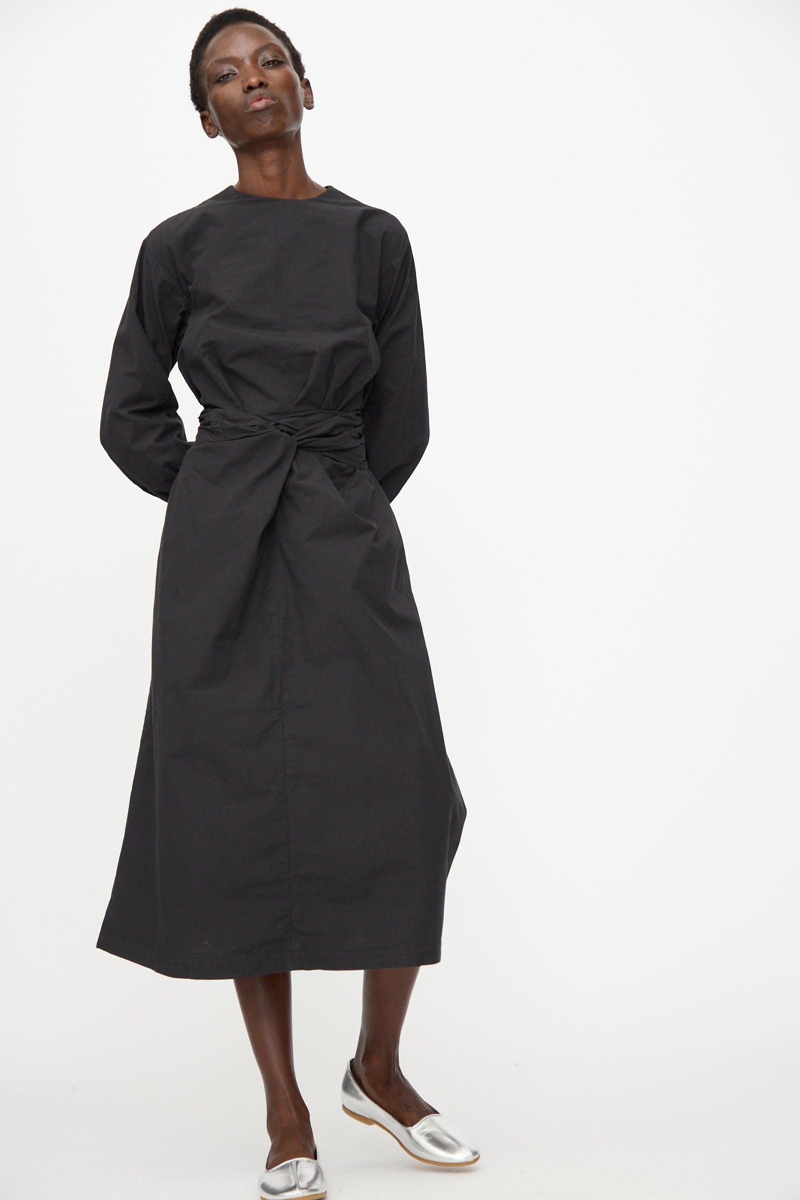A person wears the Cosmic Wonder Cotton Broadcloth Wrapped Dress in black, featuring long sleeves and a cinched waist, paired with silver flats against a plain white background.