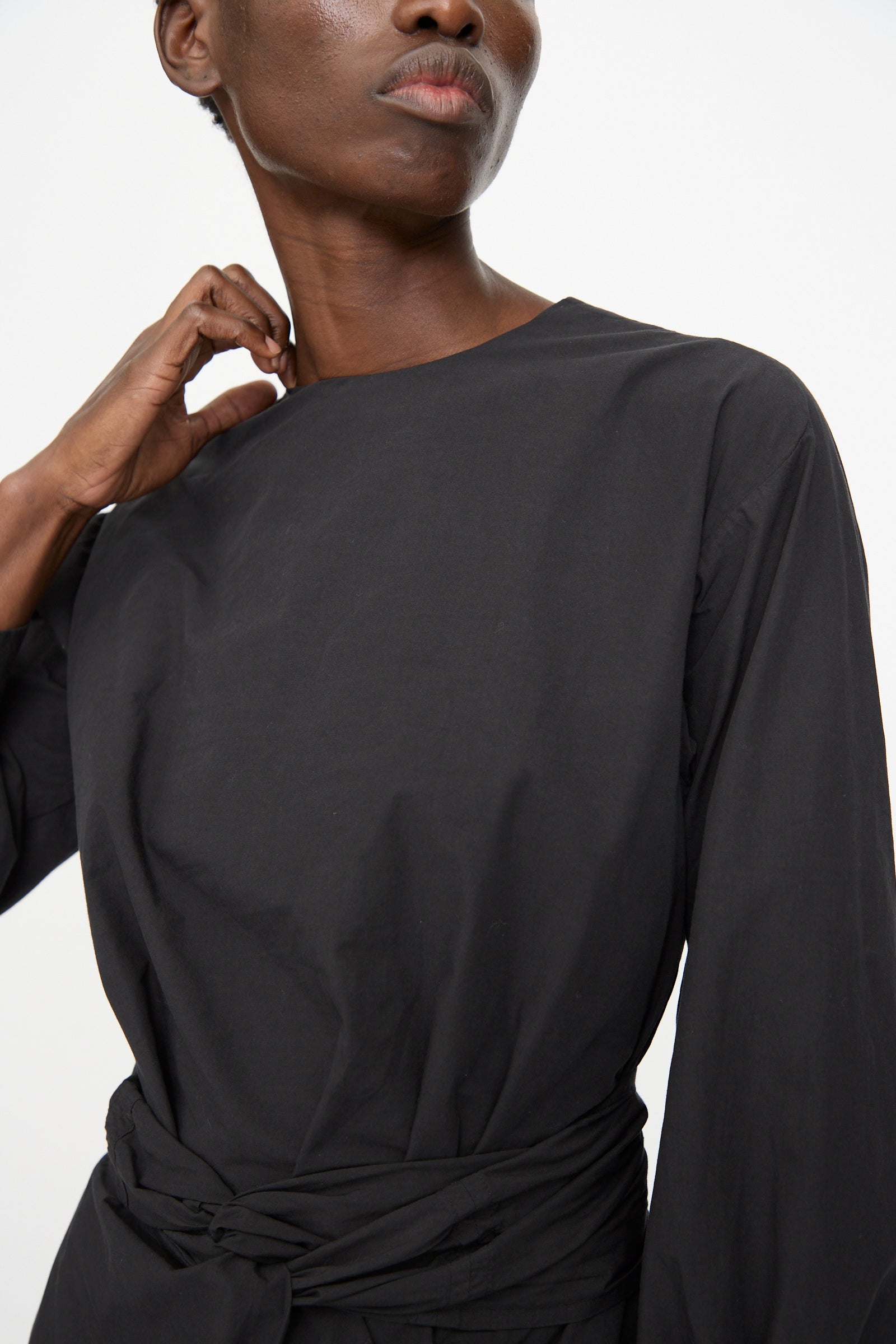 A person wearing the Cosmic Wonder Cotton Broadcloth Wrapped Dress in black stands against a plain background, one hand touching their neck.