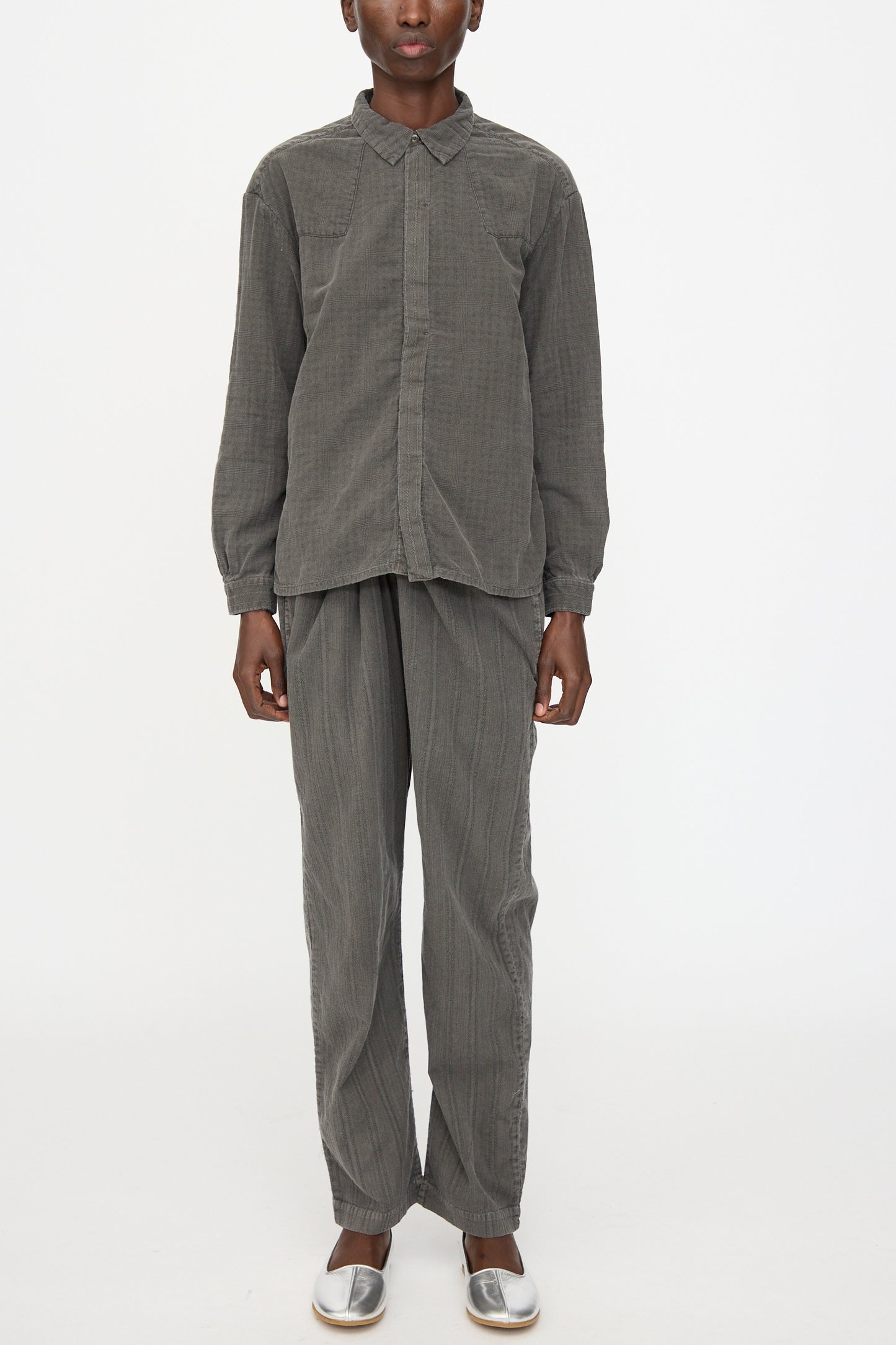 A person stands facing forward, wearing the Cosmic Wonder Double Gauze Check Country Shirt in Dark Sumi with matching gray pants and silver slip-on shoes against a plain white background.