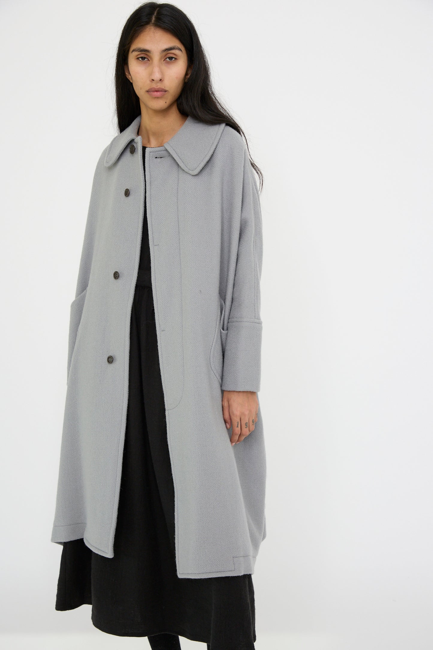 Draped over a black dress, an individual stands against a pristine white background wearing the oversized Herringbone Shetland Wool Dolman Sleeve Coat in gray from Cosmic Wonder.