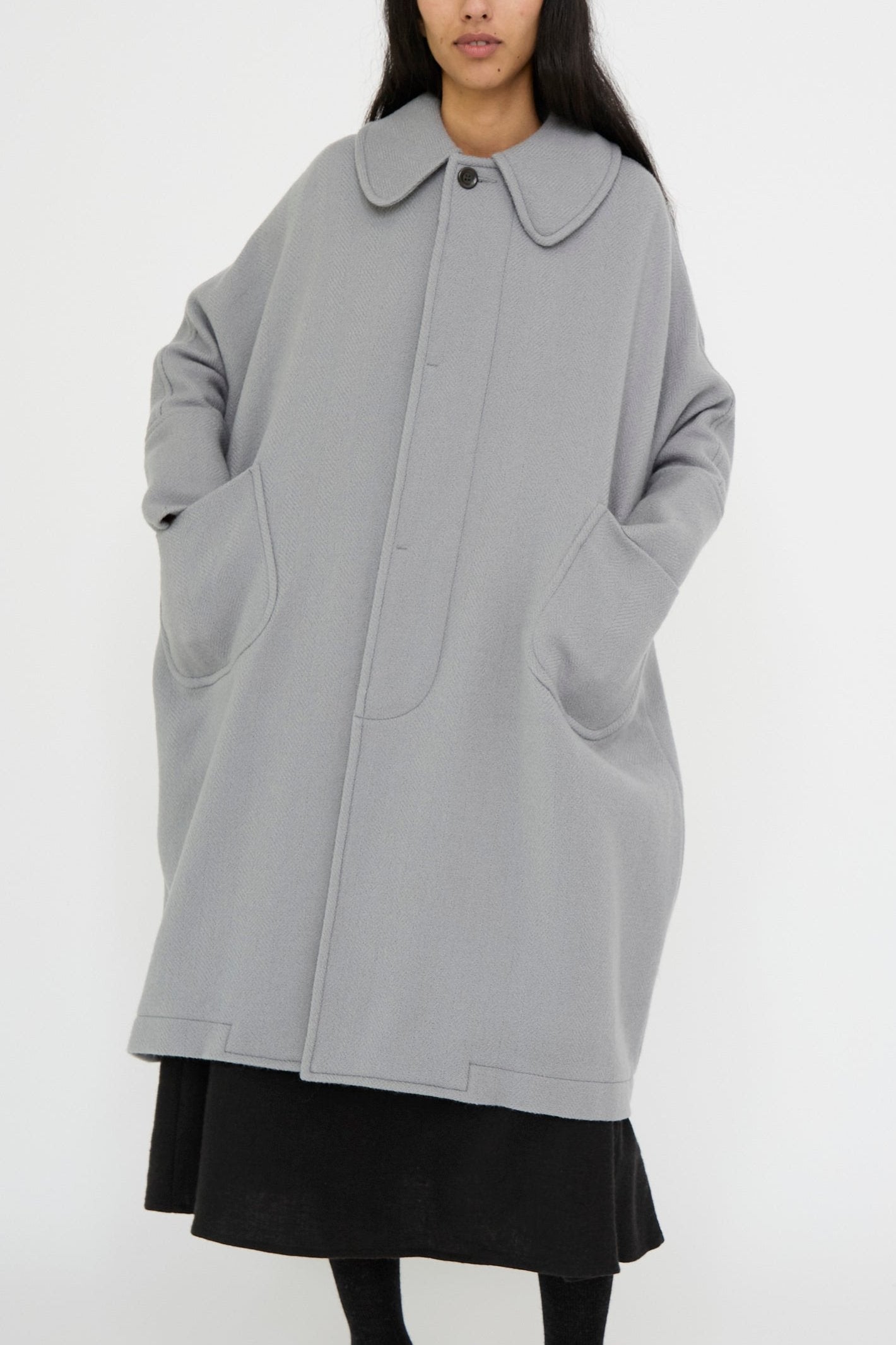 A person is standing against a plain background, wearing the Cosmic Wonder Herringbone Shetland Wool Dolman Sleeve Coat in Gray, characterized by its long, oversized design and large pockets.