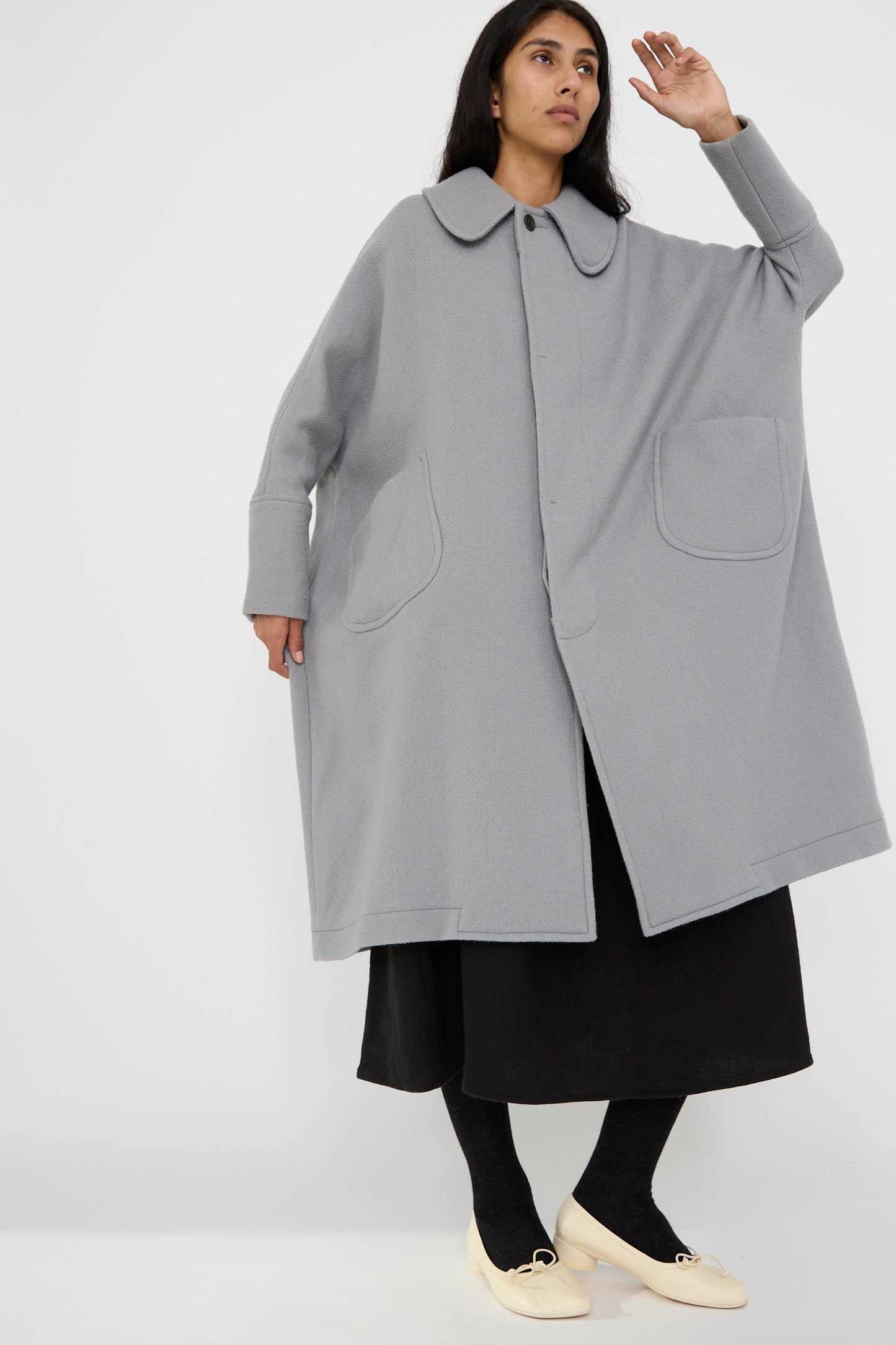 A person is dressed in a Cosmic Wonder Herringbone Shetland Wool Dolman Sleeve Coat in gray, complete with large pockets, paired with a black skirt, black tights, and cream shoes against a plain white background.