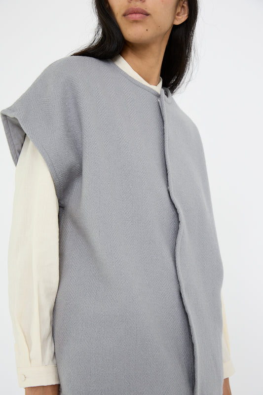 A person stands against a plain white background, wearing the Herringbone Shetland Wool Dolman Sleeve Vest in Gray by Cosmic Wonder, layered over a long-sleeve cream shirt.