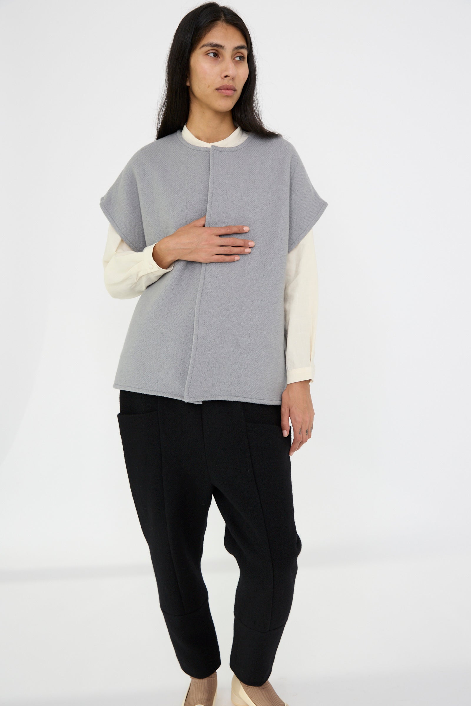 A person wearing the Herringbone Shetland Wool Dolman Sleeve Vest in Gray by Cosmic Wonder, paired with a gray sleeveless top over a white shirt and black pants, stands against a plain background, looking to the side.