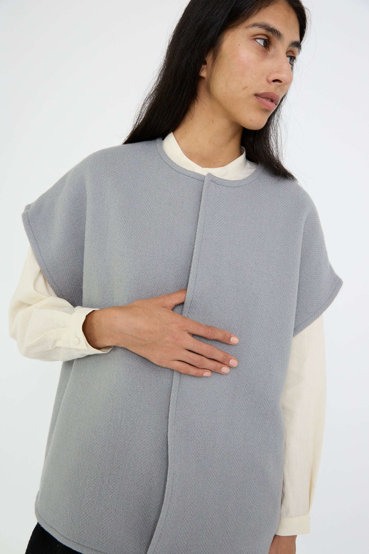 A person wearing a Cosmic Wonder Herringbone Shetland Wool Dolman Sleeve Vest in gray over a cream long-sleeve shirt, looking to the side with one hand on their chest.