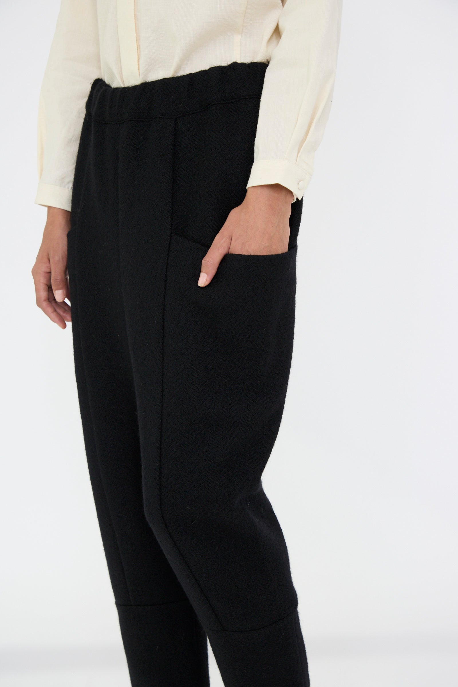 A person dressed in a cream-colored shirt and Cosmic Wonder's Herringbone Shetland Wool Winter Folk Pant in Black, featuring an elasticated waist, stands against a neutral background.