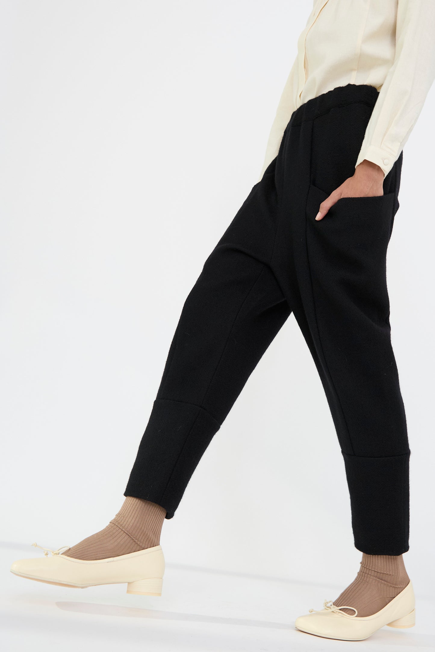 A person wearing the Herringbone Shetland Wool Winter folk Pant in Black by Cosmic Wonder, paired with a light-colored shirt, beige socks, and light shoes adorned with bows is walking against a plain white background.