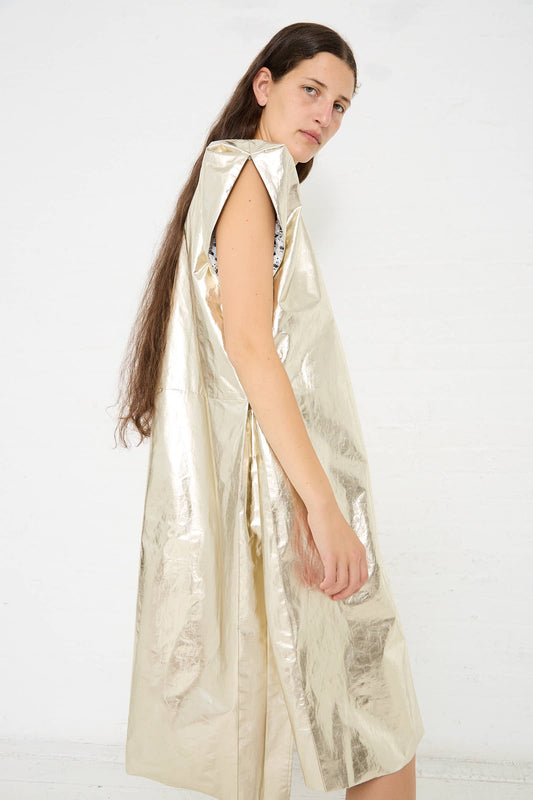 A person with long hair stands against a white background, wearing a loose, shiny, Light Foil Old Owlish Floral Paper Bag Dress by Cosmic Wonder.