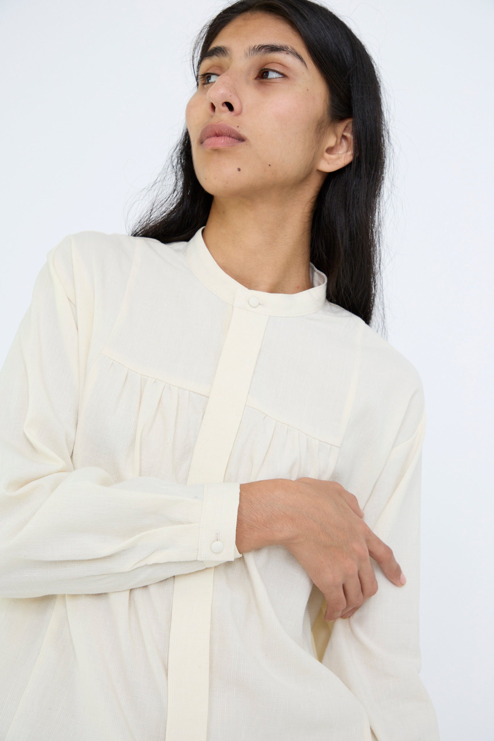 A person wearing the Cosmic Wonder Light Merino, Linen, and Cotton Glencheck Elegant Gathered Shirt in white with long sleeves, button details, and a band collar looks to the side against a plain background.