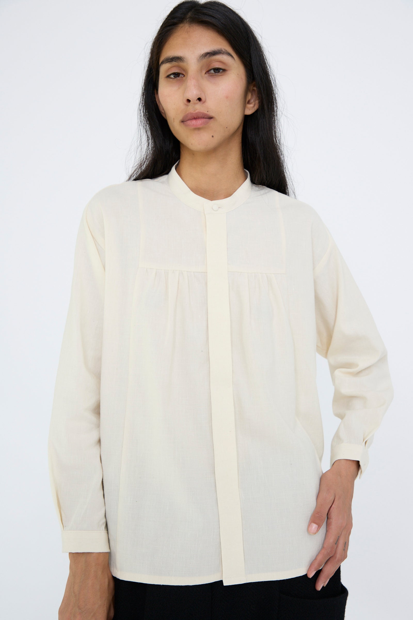 Against a plain background, a person with long hair is wearing the Cosmic Wonder Light Merino, Linen and Cotton Glencheck Elegant Gathered Shirt in White, featuring long sleeves and a band collar.