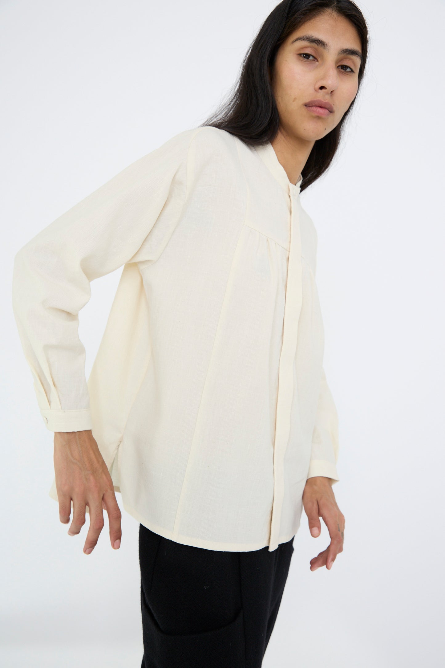 A person with long hair wearing a white Light Merino, Linen, and Cotton Glencheck Elegant Gathered Shirt by Cosmic Wonder along with black pants stands against a white background.