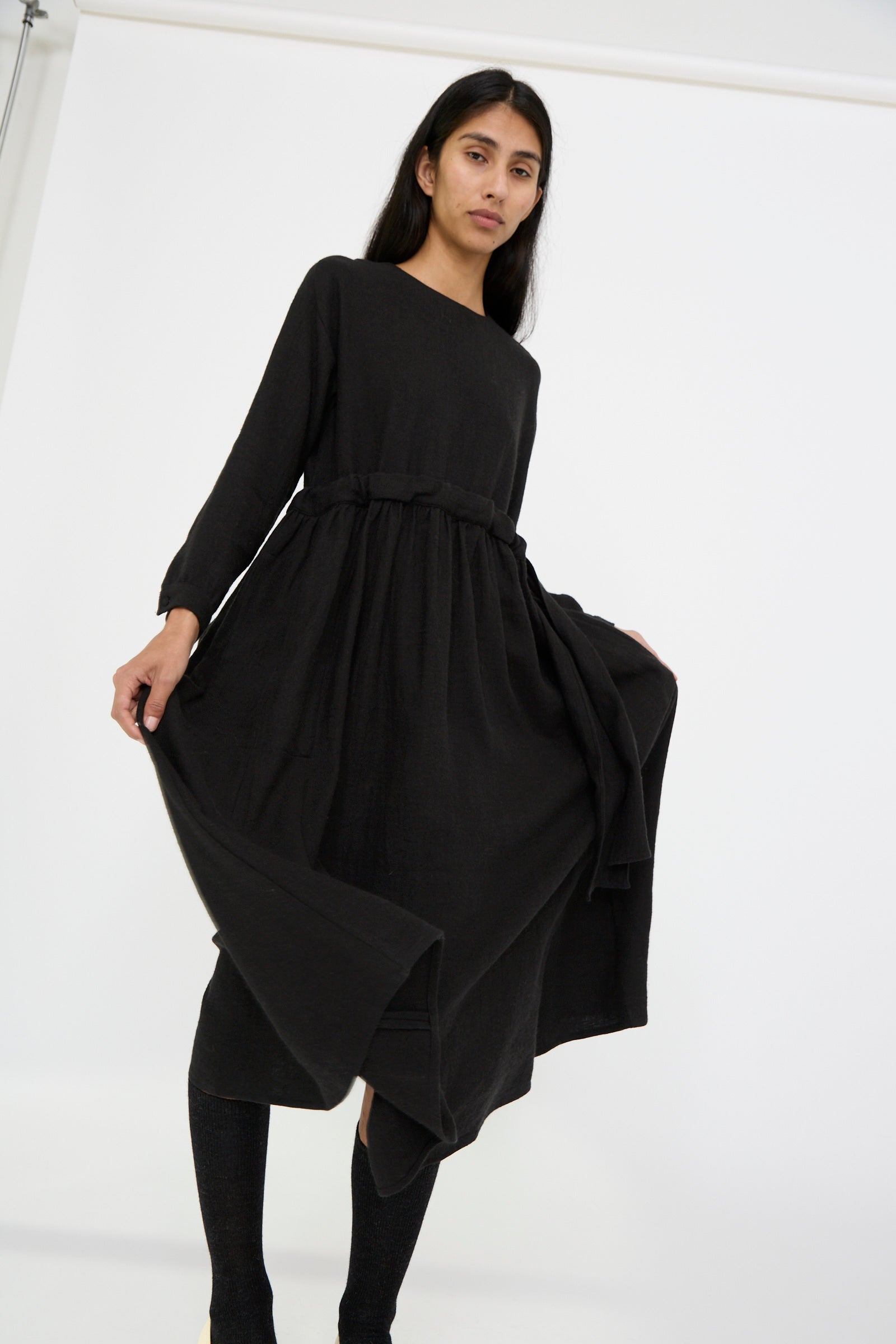 A person in the Linen and Wool Twill Old Linen Farmer Dress in Black by Cosmic Wonder, a person poses against a plain white backdrop. 