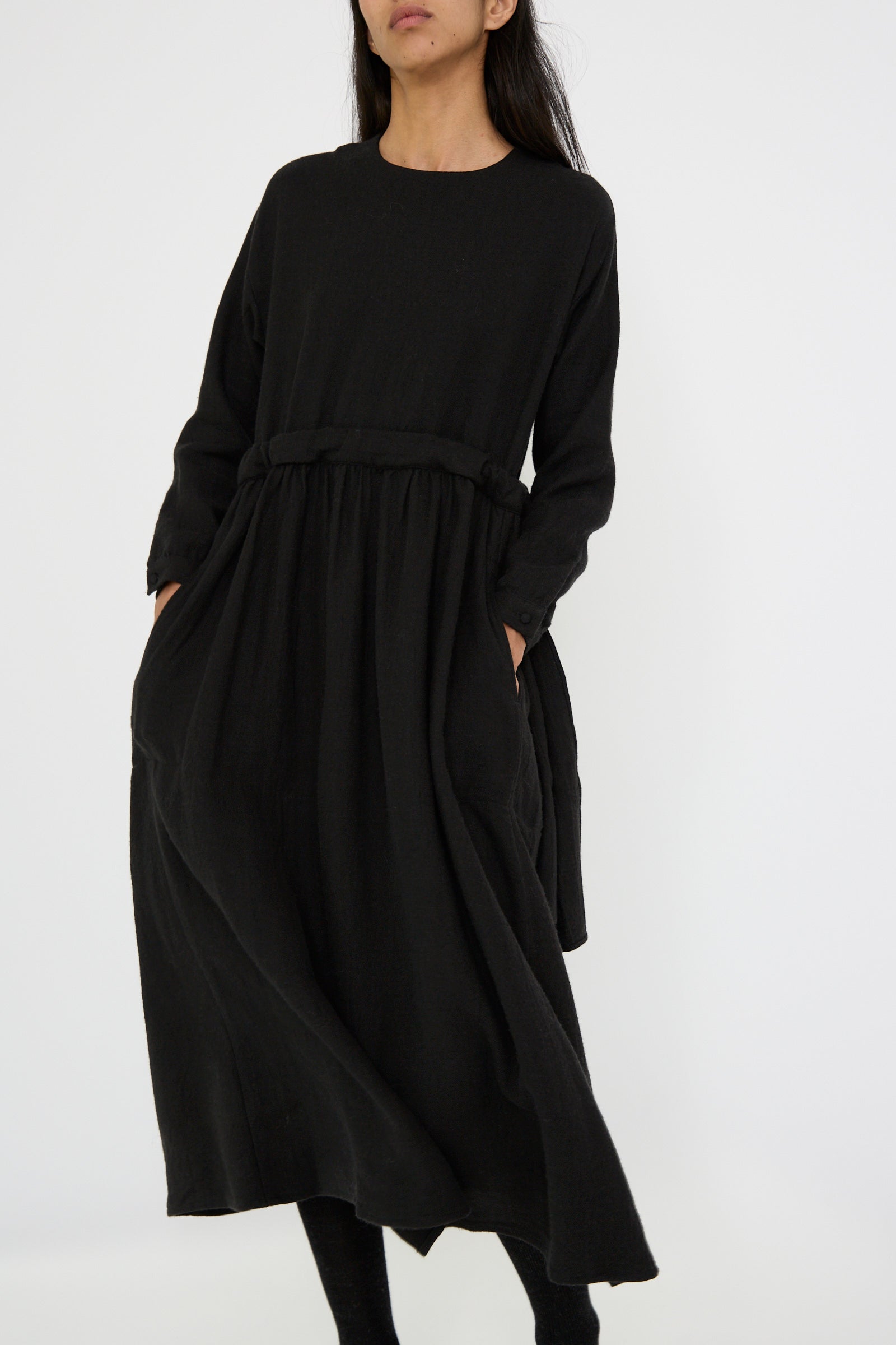 A person wearing the Cosmic Wonder Linen and Wool Twill Old Linen Farmer Dress in black, a relaxed fit maxi dress featuring long sleeves and pockets, stands against a plain white background.