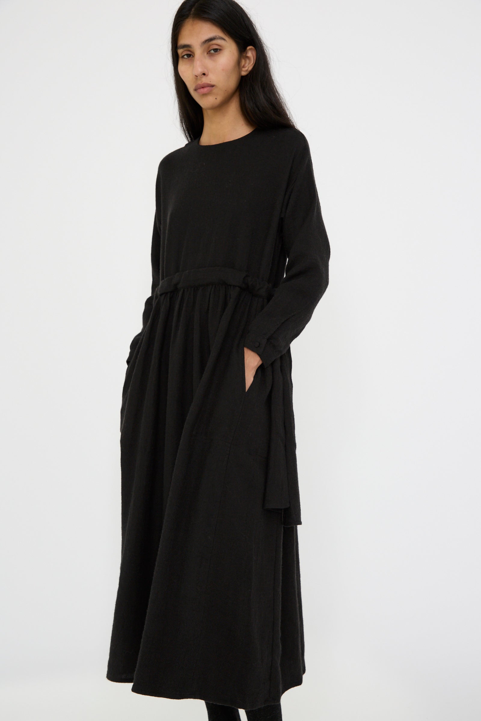 An individual is seen against a white backdrop, dressed in a relaxed-fit Linen and Wool Twill Old Linen Farmer Dress in Black with pockets and long sleeves by Cosmic Wonder.