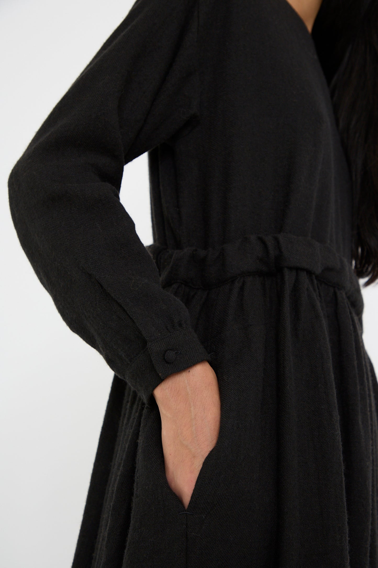 A person wearing the Linen and Wool Twill Old Linen Farmer Dress in black from Cosmic Wonder, featuring a relaxed-fit and long sleeves, with their hand in a side pocket, against a plain background.