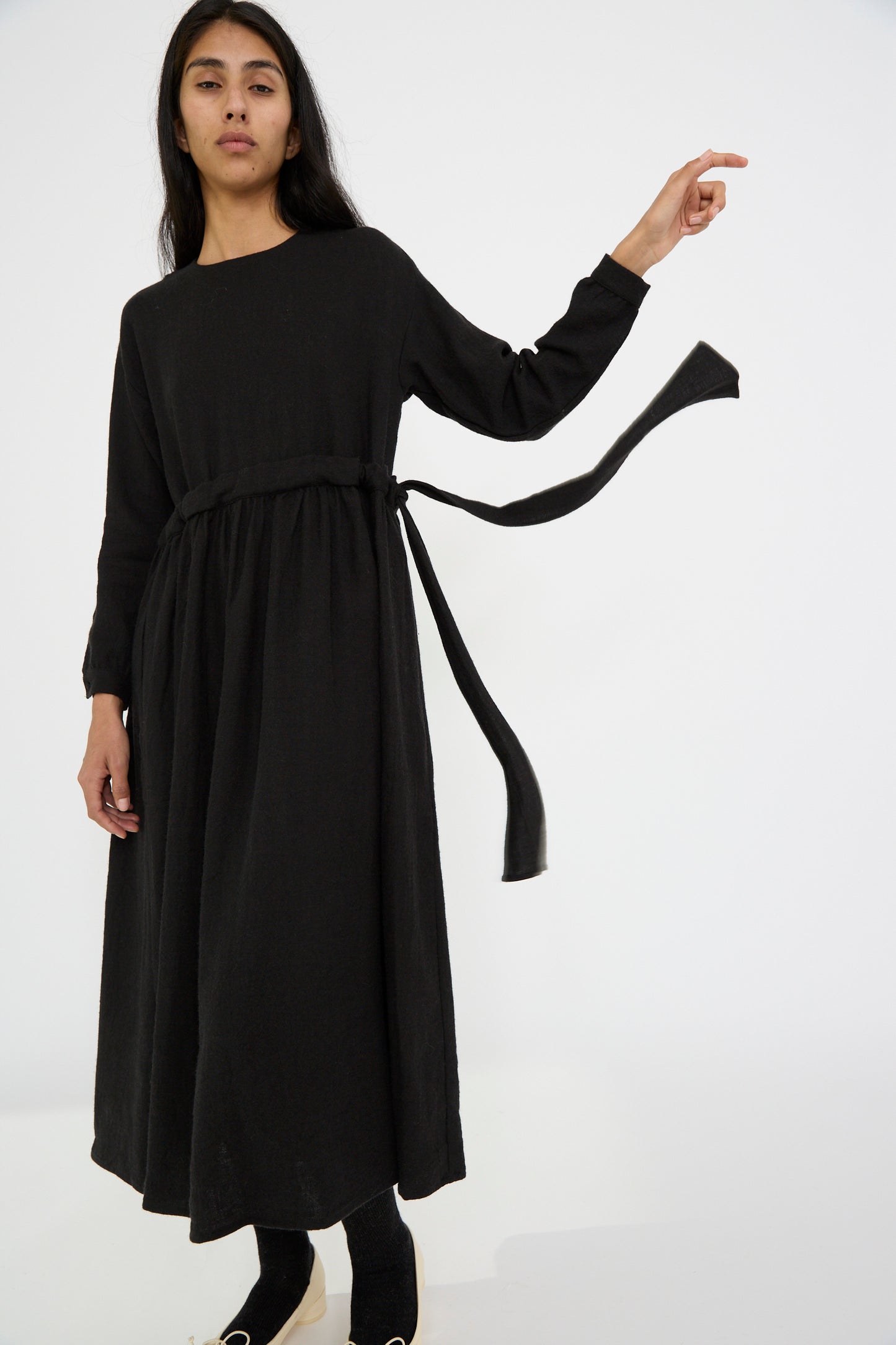 Against a plain white background, a person with one arm raised wears the Cosmic Wonder's Linen and Wool Twill Old Linen Farmer Dress in Black. The dress features a tied belt and relaxed fit, crafted from a linen wool blend fabric. 