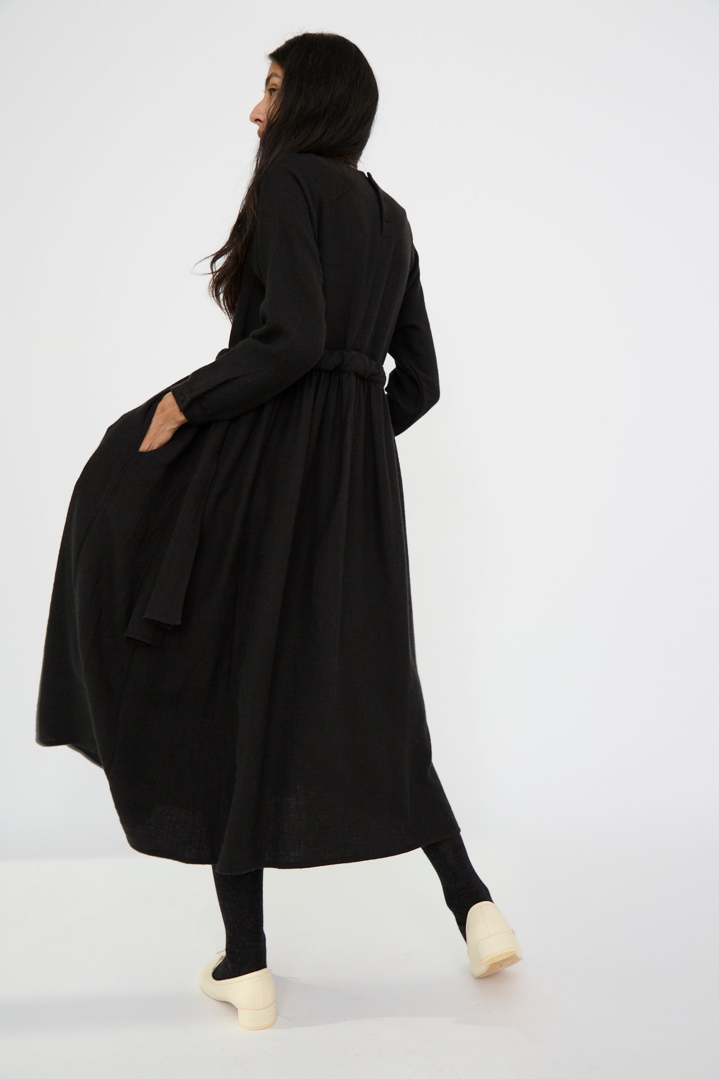 A person is seen from the back wearing a Cosmic Wonder Linen and Wool Twill Old Linen Farmer Dress in Black, paired with black leggings and beige shoes, against a plain white background.