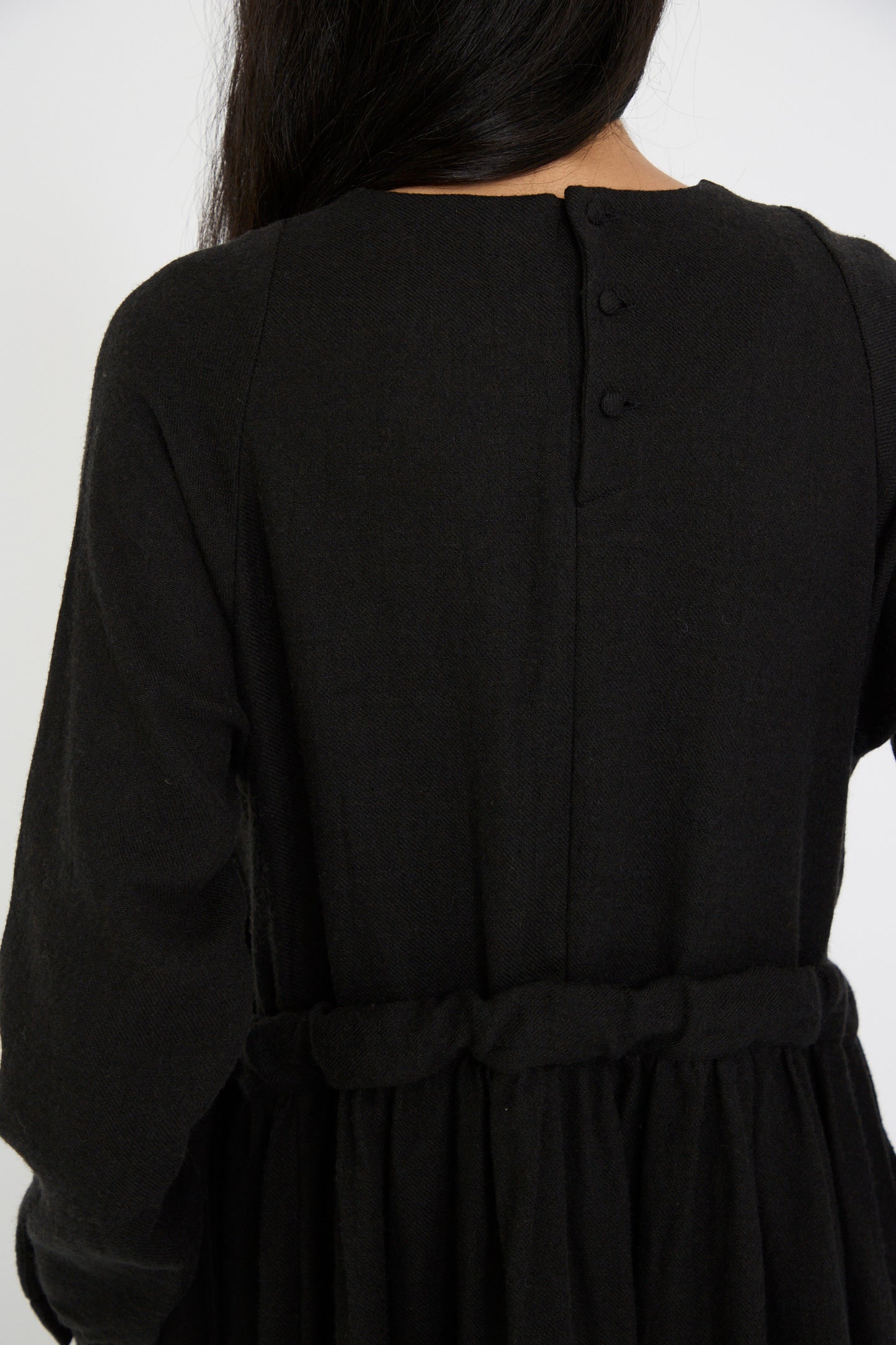 A person is shown from the back close up wearing the Cosmic Wonder "Linen and Wool Twill Old Linen Farmer Dress" in black, featuring button details and a fabric belt, crafted from a linen wool blend for a relaxed fit.