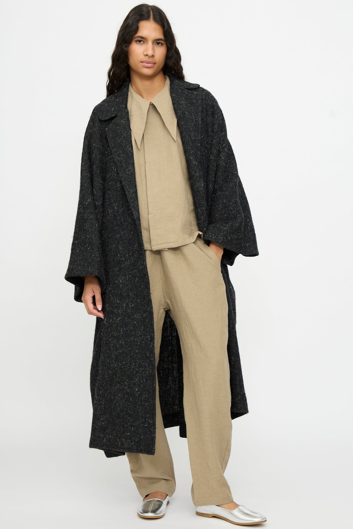 A person wears a black Cosmic Wonder Nostalgic Painter’s Winter Coat over a beige shirt and trousers, with metallic silver flat shoes.