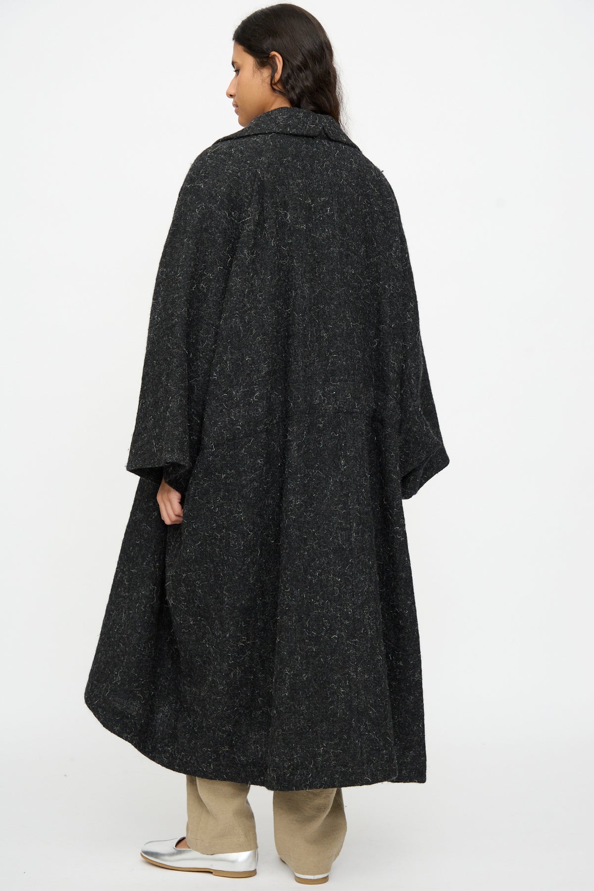 A person seen from the back wearing a Cosmic Wonder Nostalgic Painter’s Winter Coat in Black with wide sleeves over tan trousers and silver shoes.