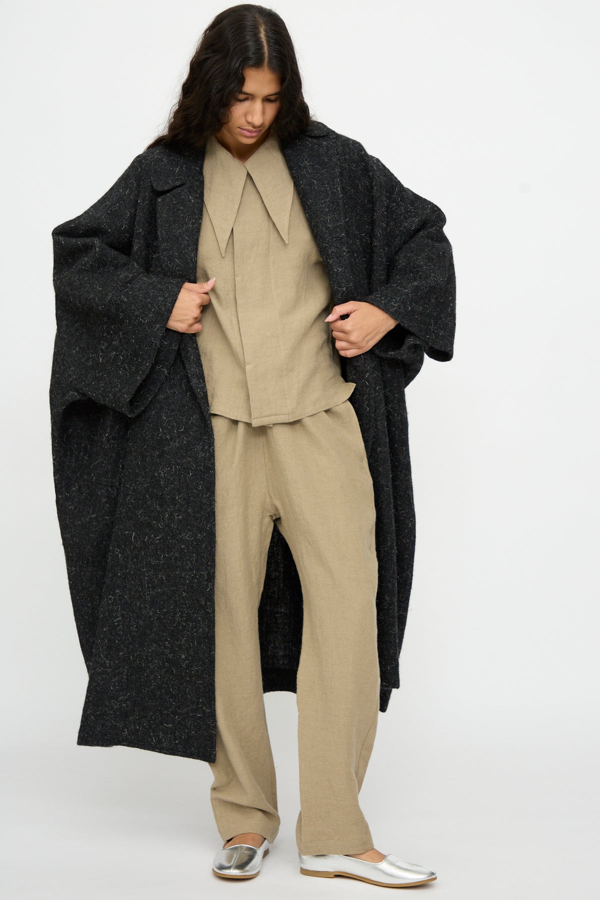 Person wearing an oversized Cosmic Wonder Nostalgic Painter’s Winter Coat in Black over a beige shirt and trousers with silver shoes.