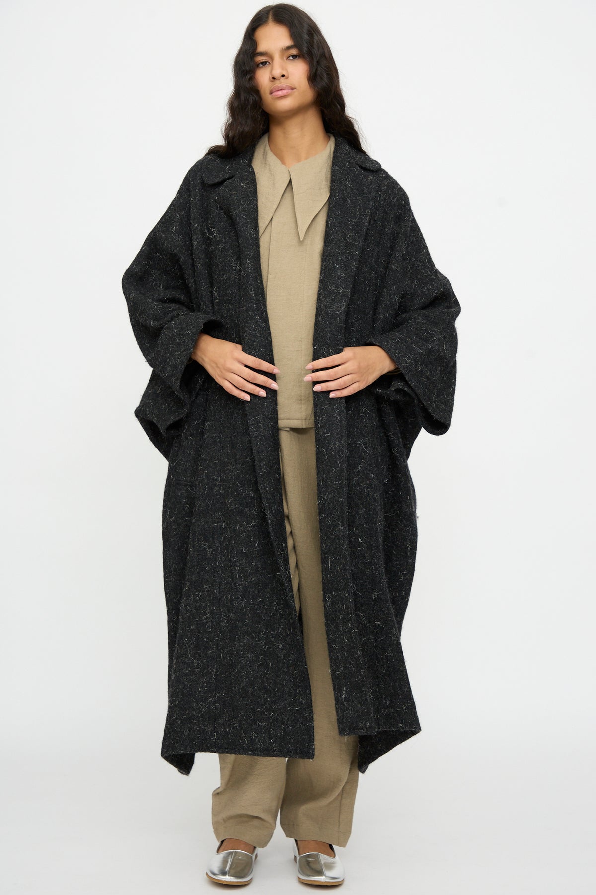 A person wears an oversized, black Nostalgic Painter’s Winter Coat by Cosmic Wonder with wide sleeves over a beige collared shirt and trousers.