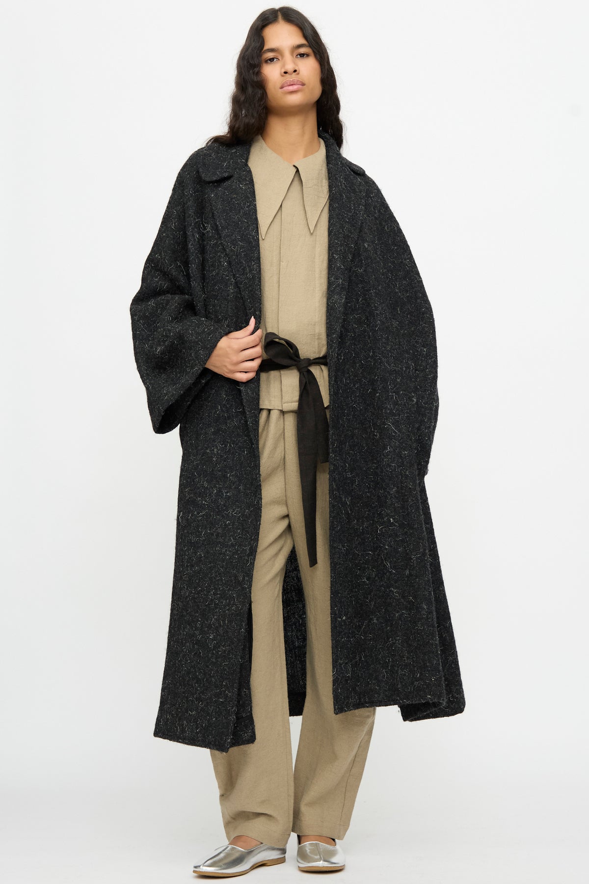Person wearing an oversized, black textured Cosmic Wonder Nostalgic Painter’s Winter Coat in Black over a beige collared jumpsuit tied with a black sash.
