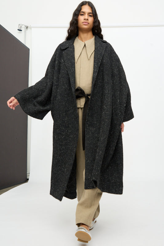 A person wears a long, oversized black Cosmic Wonder Nostalgic Painter’s Winter Coat with wide sleeves over a beige collared outfit.