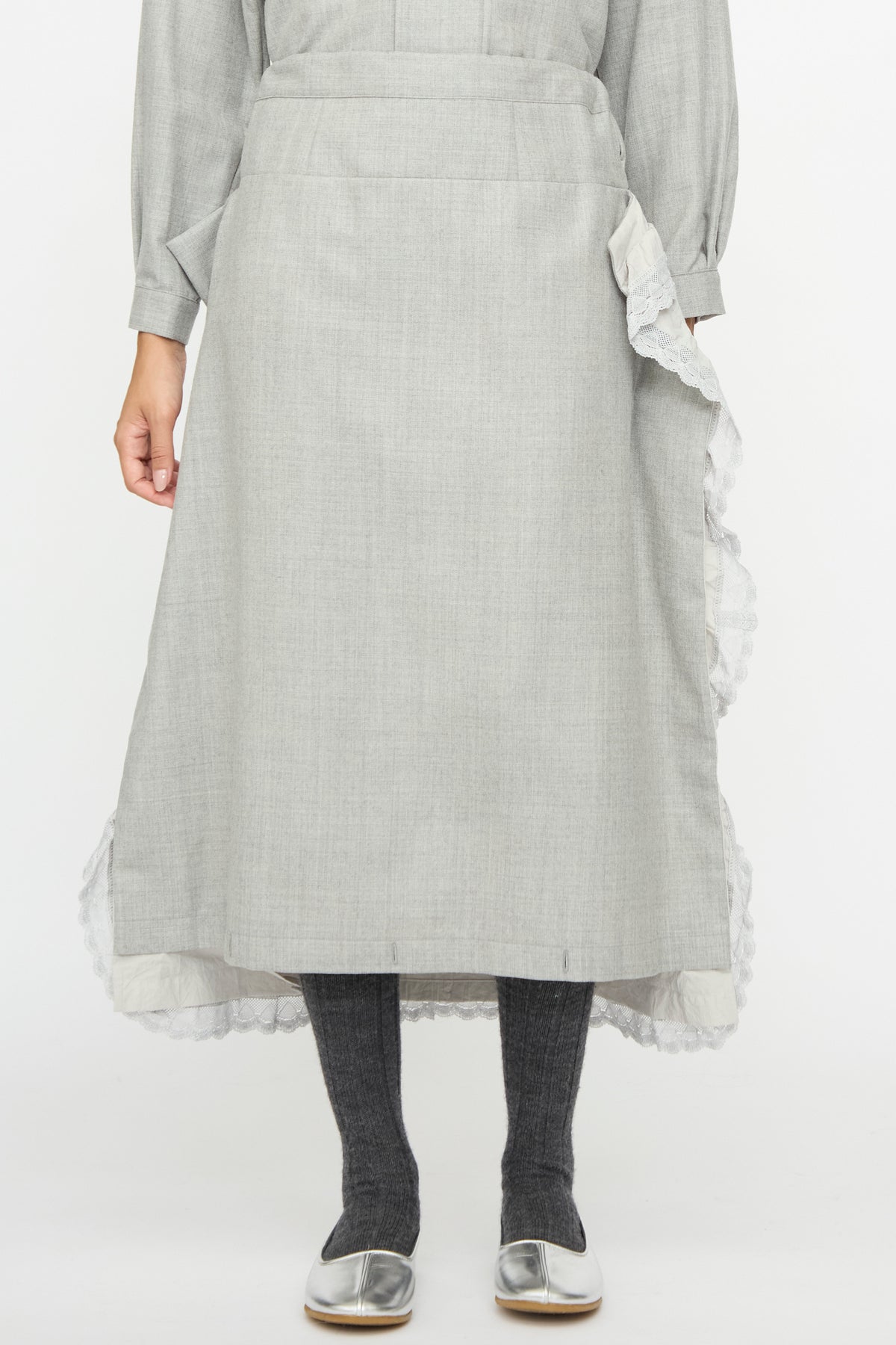 Cosmic Wonder Nostalgic Painter’s Work Skirt in gray, a midi length made in Japan featuring an asymmetrical, ruffled lace trim along the side, worn with dark gray tights and silver flats.