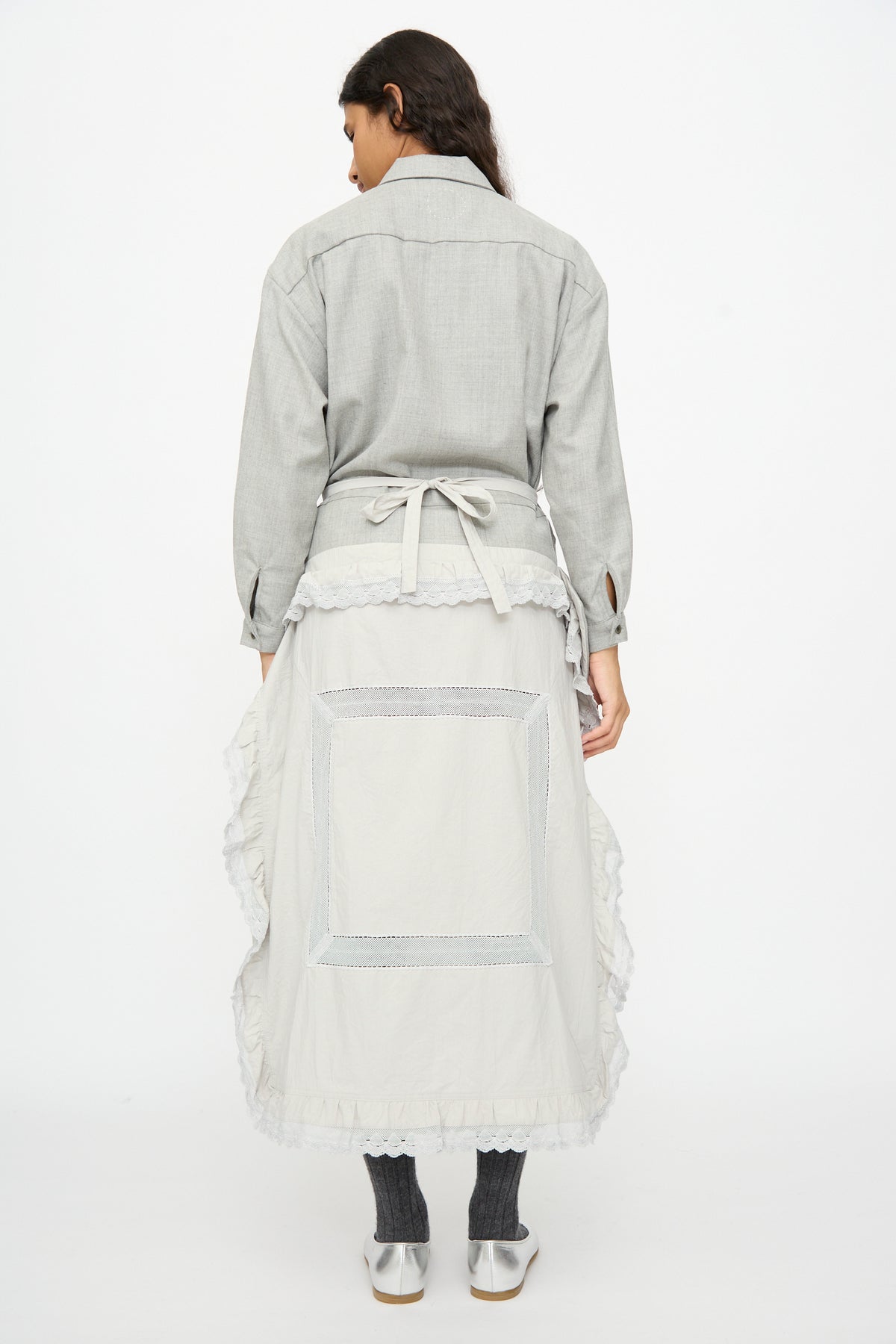A person from behind wears a gray button-down shirt and a Cosmic Wonder Nostalgic Painter’s Work Skirt in Gray with a square panel, ruffled trim, and tie-waist, paired with dark leggings and silver shoes.