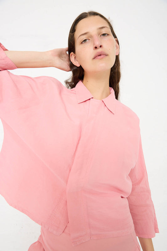 Person with long hair, wearing a loose-fitting Silk Paper Dolman Sleeve Shirt in Peach Jade by Cosmic Wonder, stands against a plain white background, with their right hand resting on the back of their head.