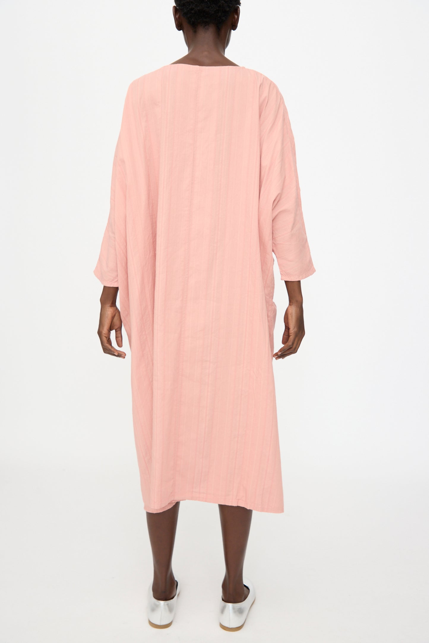 A person faces away from the camera against a white background, wearing Cosmic Wonder’s Stripe Combination Twisted Dress in Rose Crystal with long sleeves and white slip-on shoes.