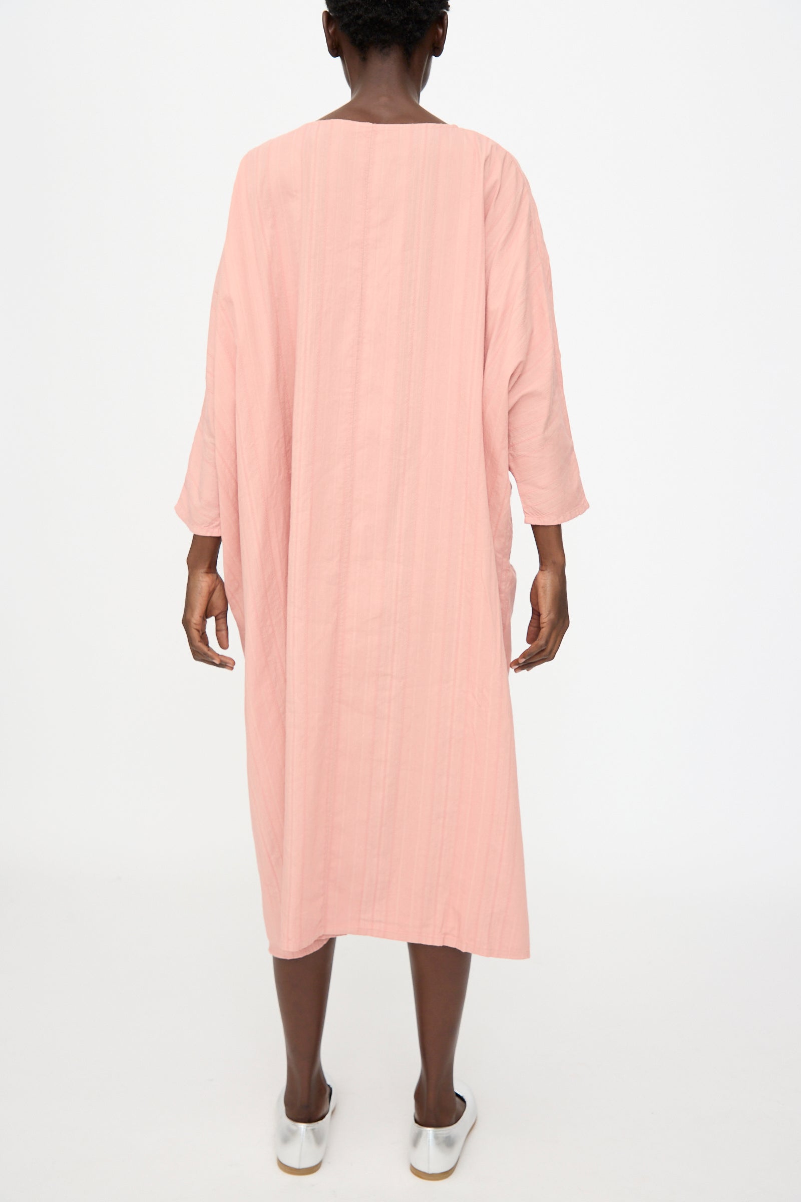 A person faces away from the camera against a white background, wearing Cosmic Wonder’s Stripe Combination Twisted Dress in Rose Crystal with long sleeves and white slip-on shoes.